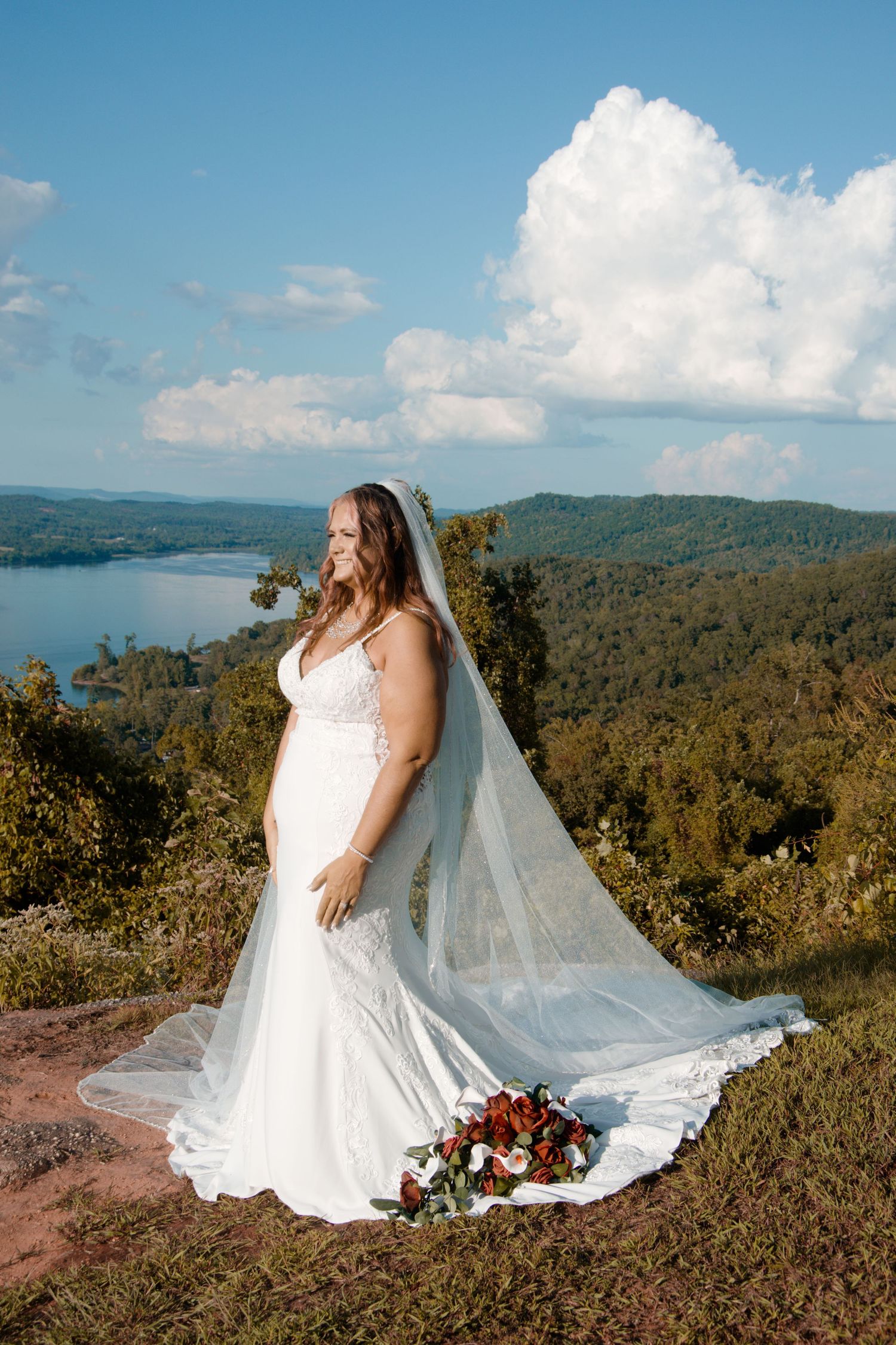 A bride in a white lace gown with flowing veil poses on a mountaintop overlooking a scenic lake view.