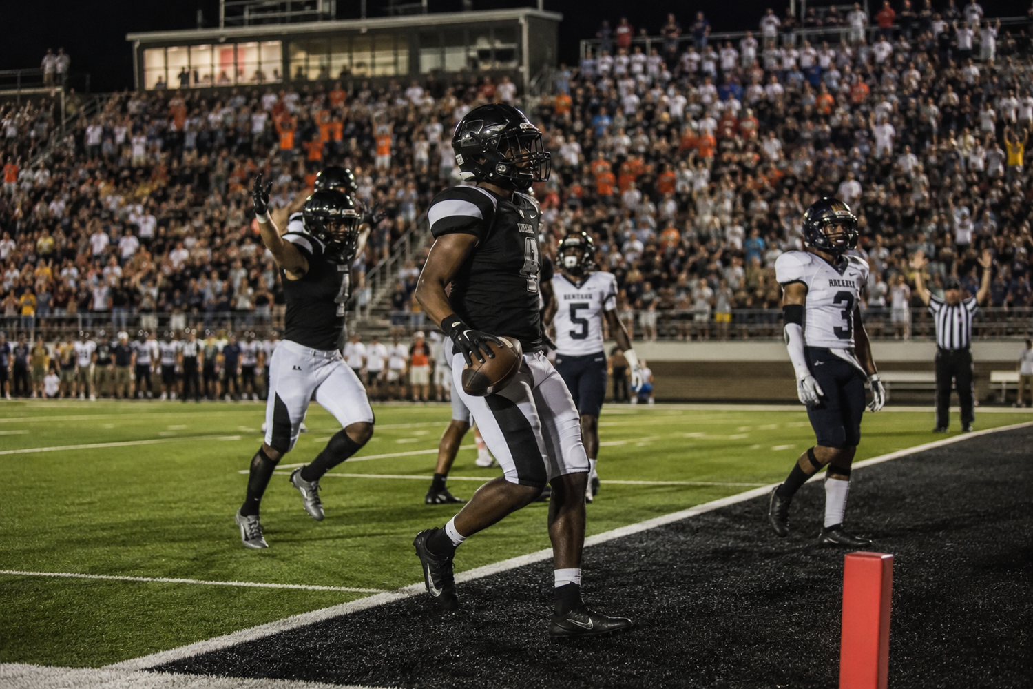 Football player scores a touchdown in an end zone during a night game under bright stadium lights.