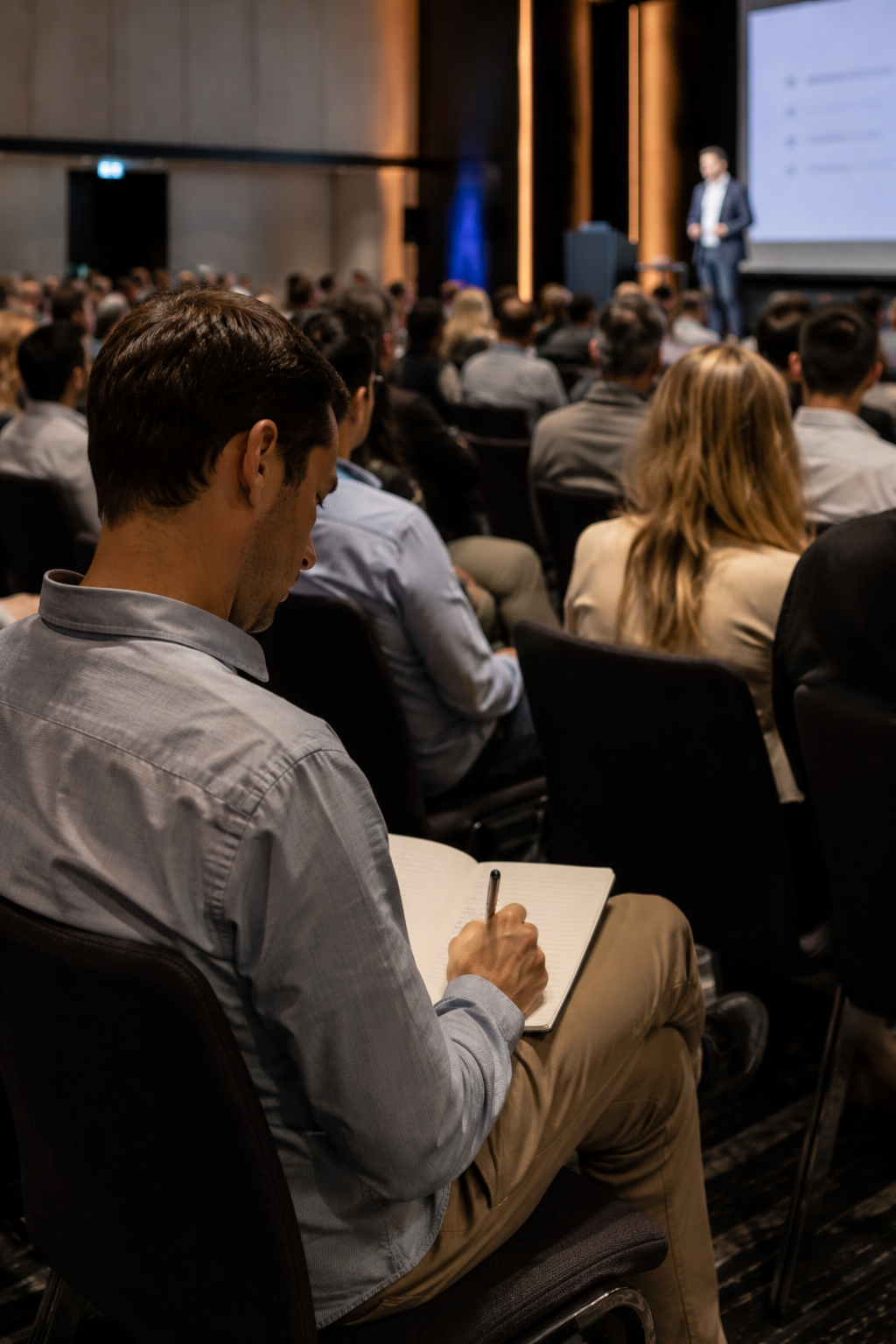 Businessman taking notes in notebook while attending a professional conference with speaker on stage.