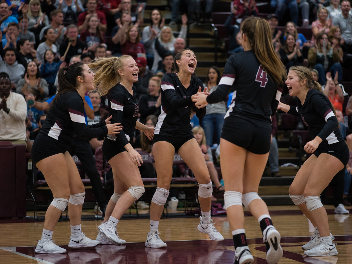 Excited volleyball team in black uniforms celebrates a point victory together on a crowded gymnasium court.