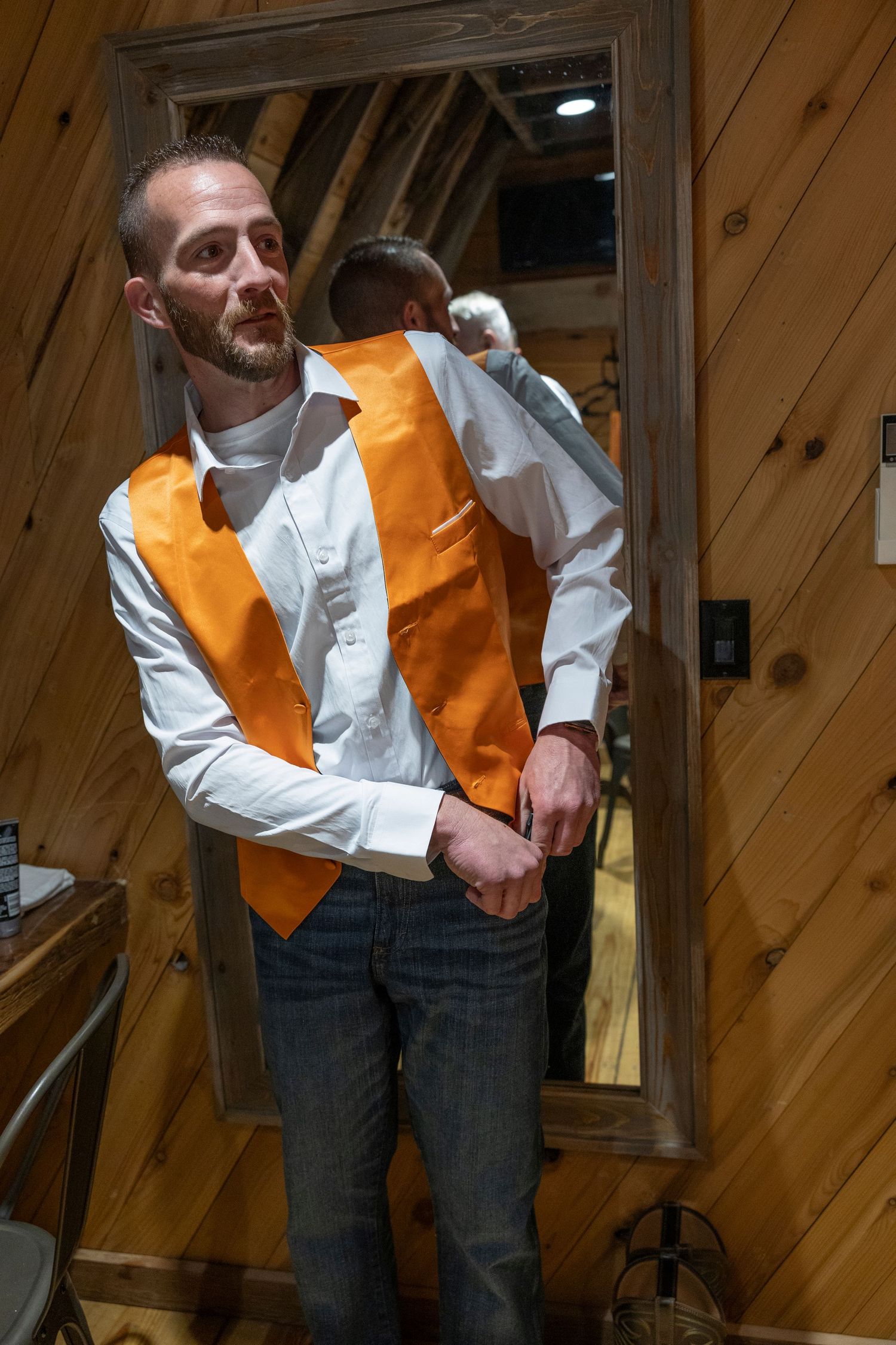 Man in white shirt and orange vest adjusts his outfit while standing before a mirror in a wooden cabin room.