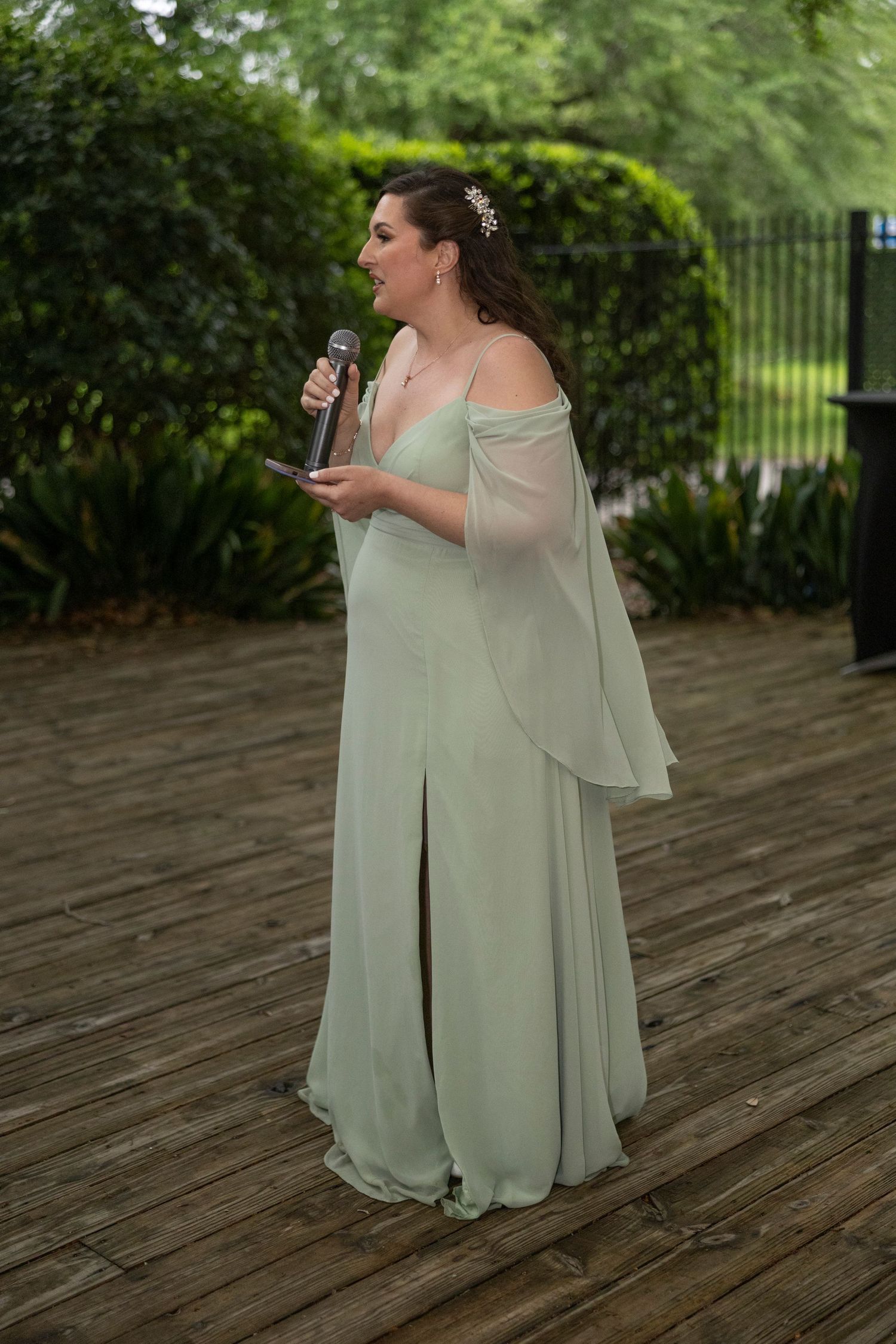 A person in a flowy sage green bridesmaid dress with off-shoulder sleeves holds a drink while standing on a wooden deck.