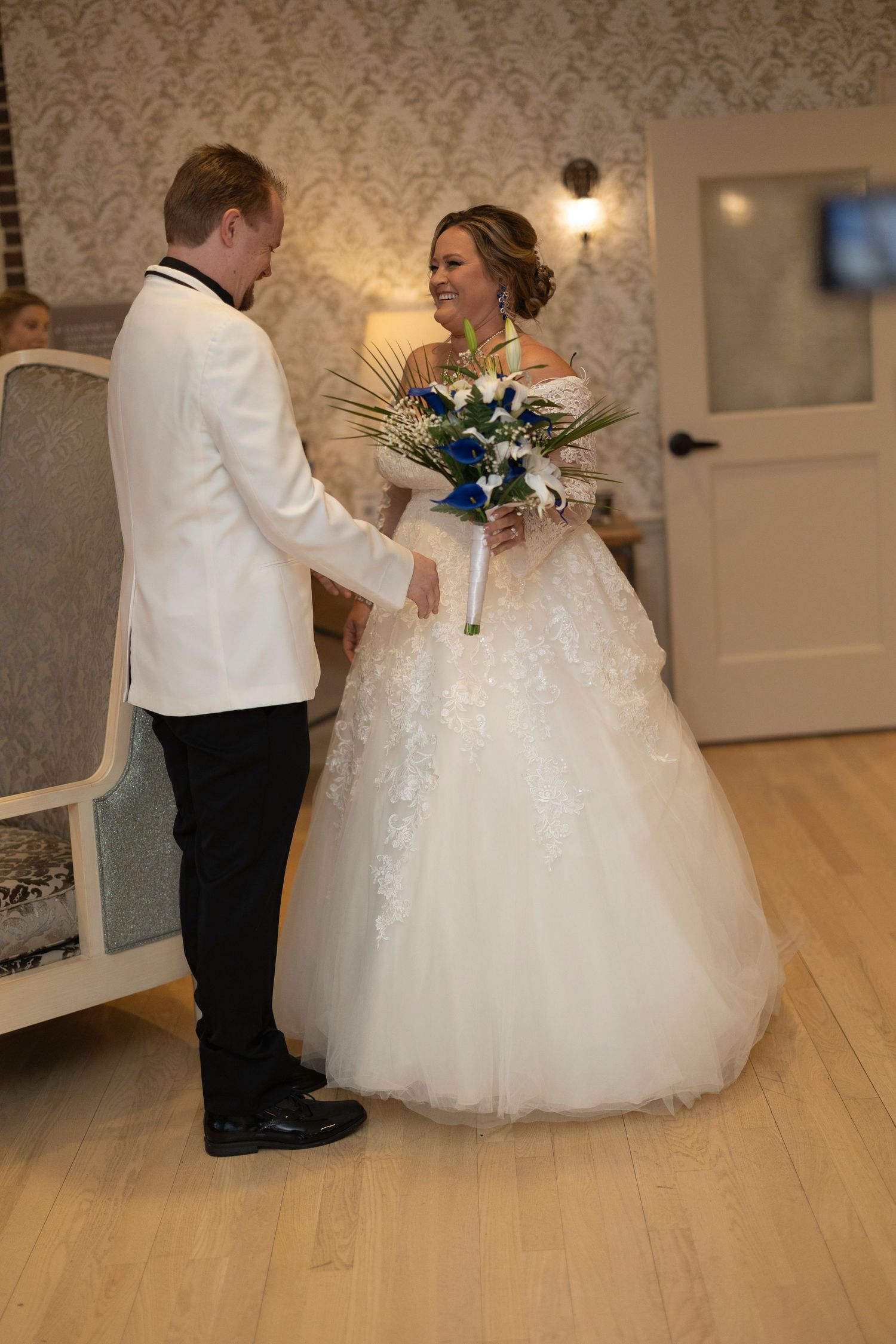 Bride in white ball gown holding blue and white bouquet stands with groom in white jacket at indoor wedding venue.