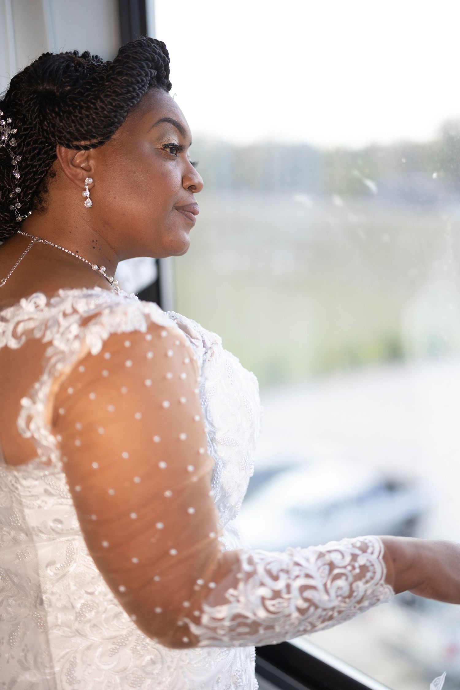 Bride in lace long-sleeve wedding dress gazes out window, wearing pearl earrings and necklace with braided updo.