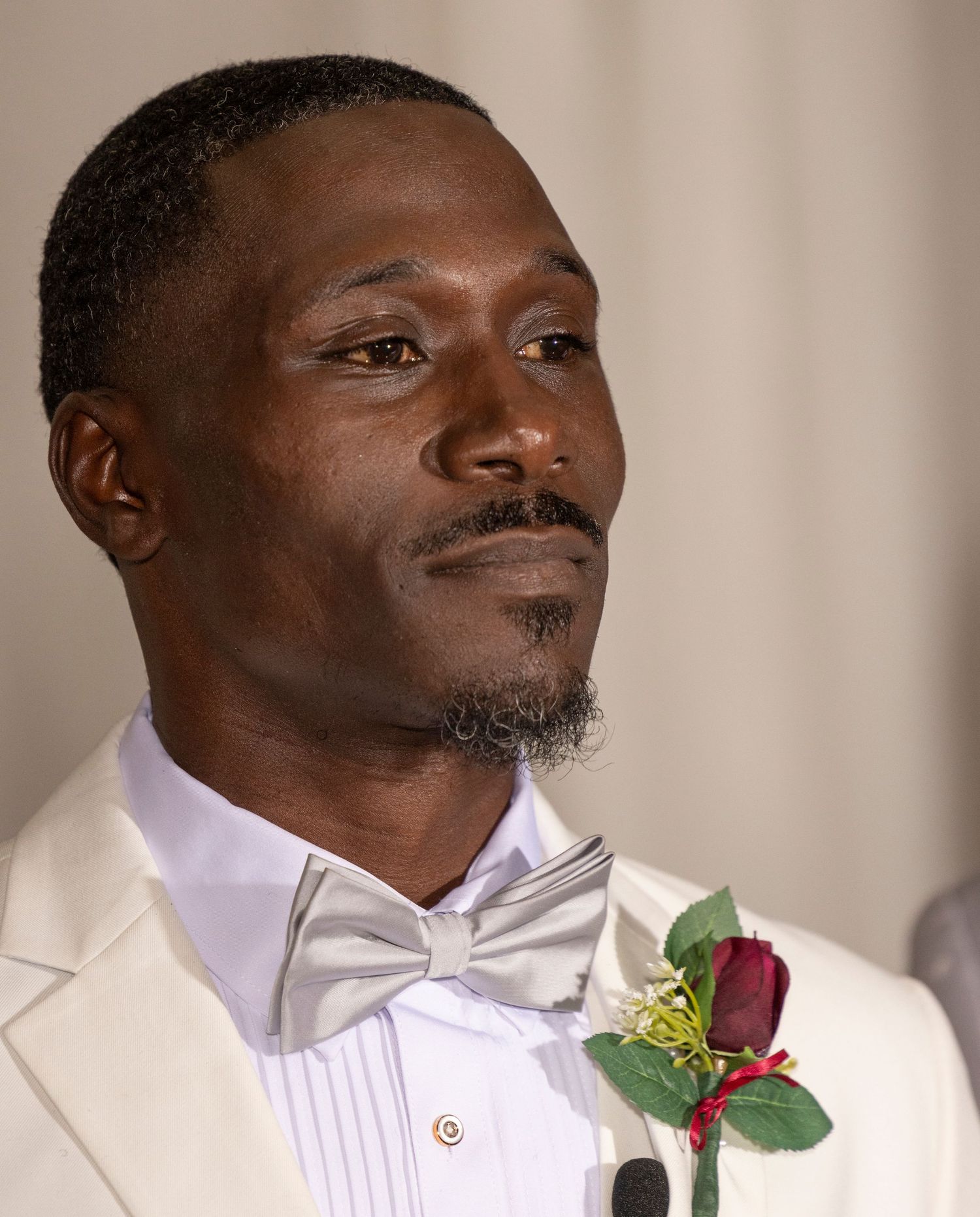 Man in white tuxedo with bow tie and red rose boutonniere at formal event.