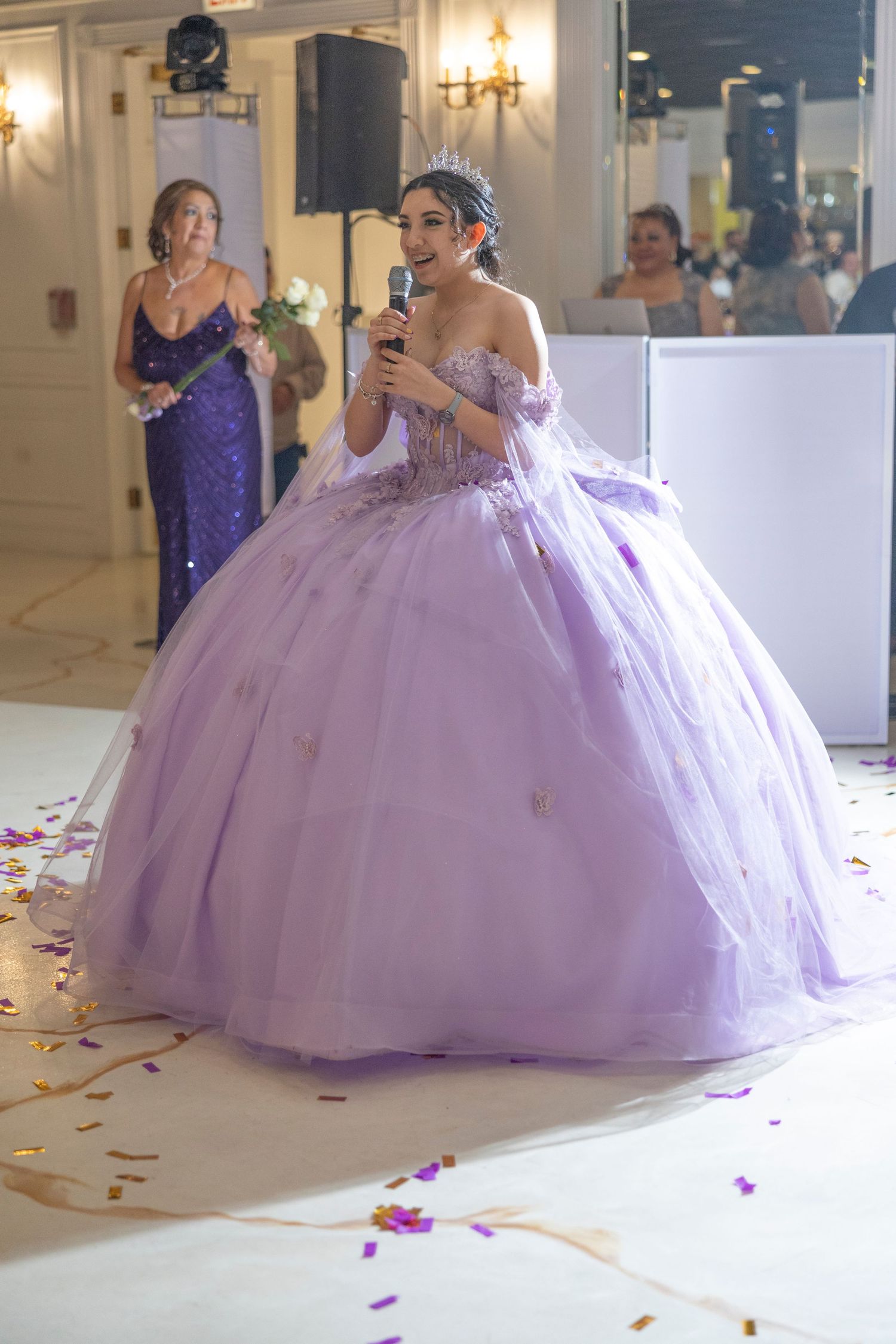 A person in a lavender ball gown stands in an elegant reception hall with rose petals scattered on the floor.