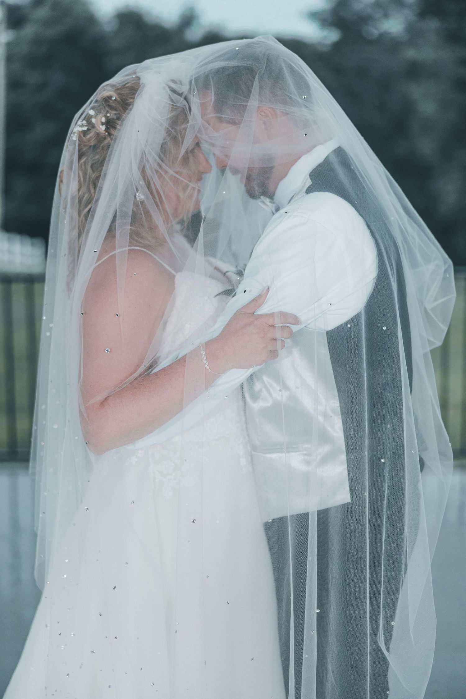 A romantic wedding couple shares an intimate kiss under a flowing white veil in an outdoor setting.