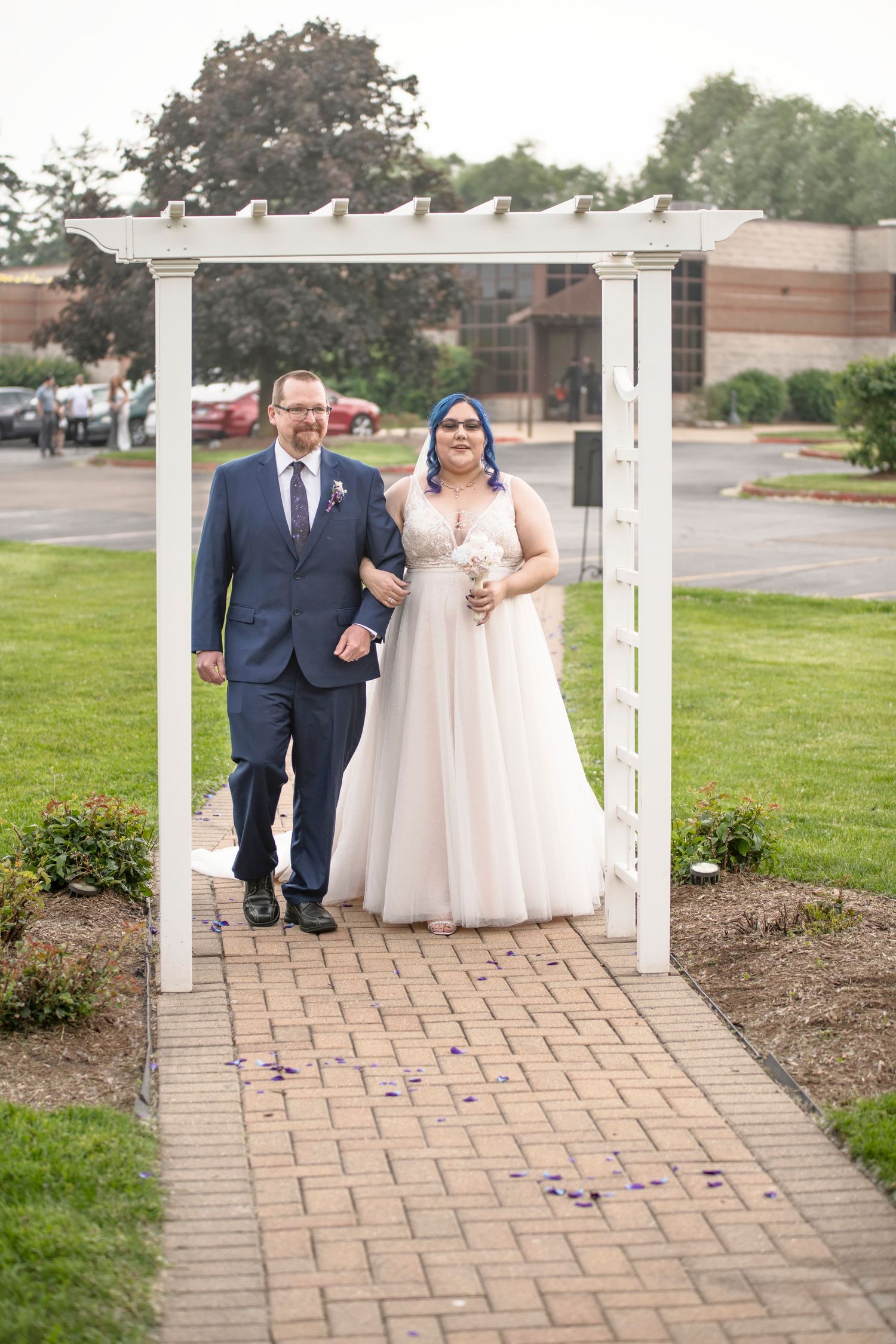 A couple in formal wedding attire walks down a stone paved path framed by white pillars at an outdoor venue.