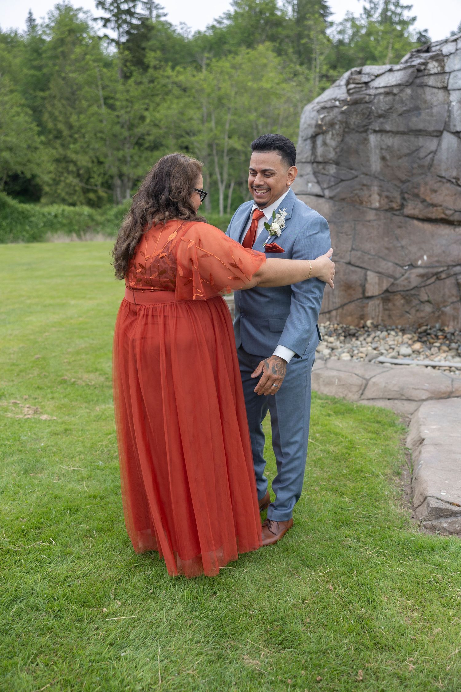 A couple in a blue suit and rust-colored dress dance playfully on green grass near a stone wall.