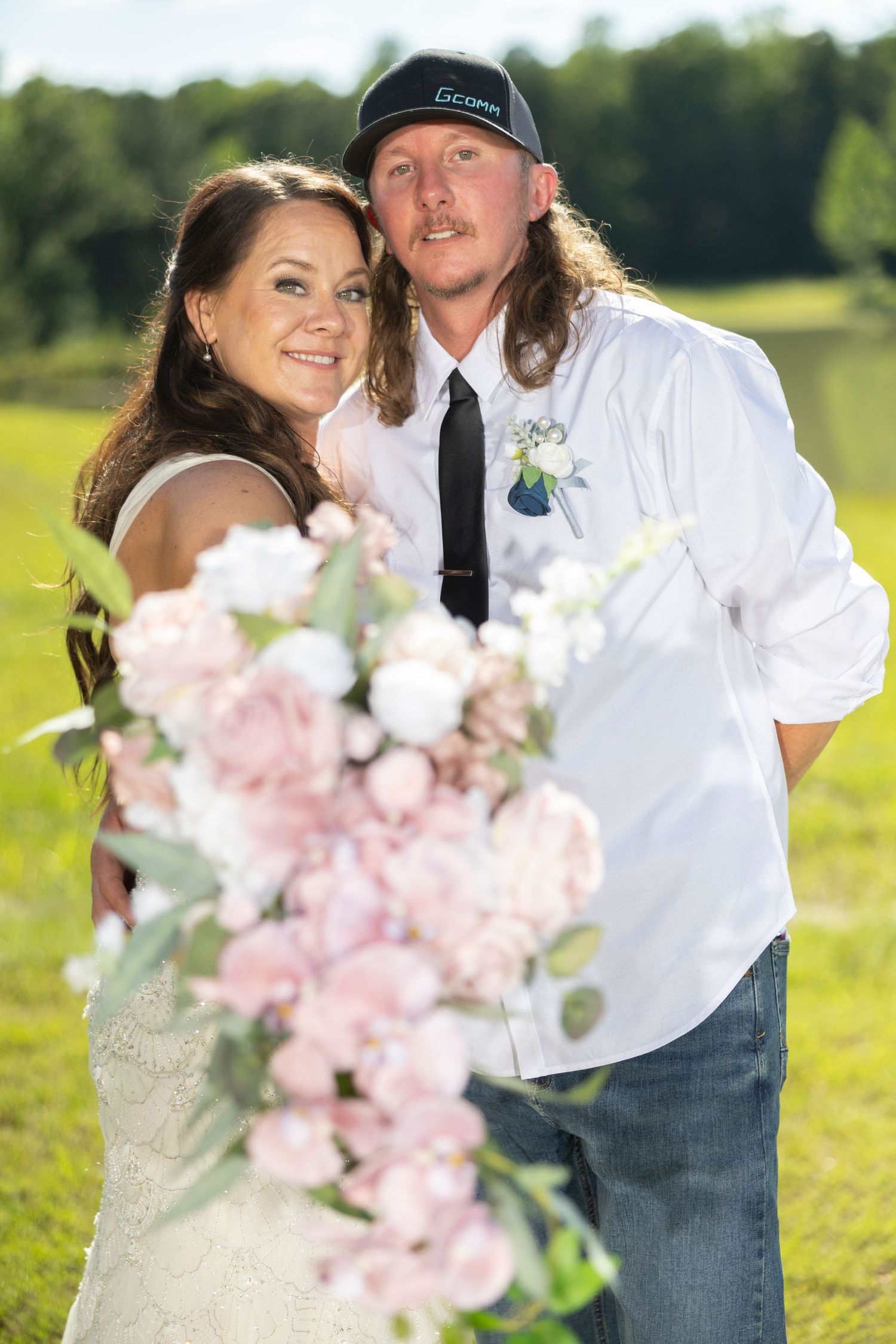 A couple in wedding attire poses together in a sunny field with pink and white flowers in the foreground.