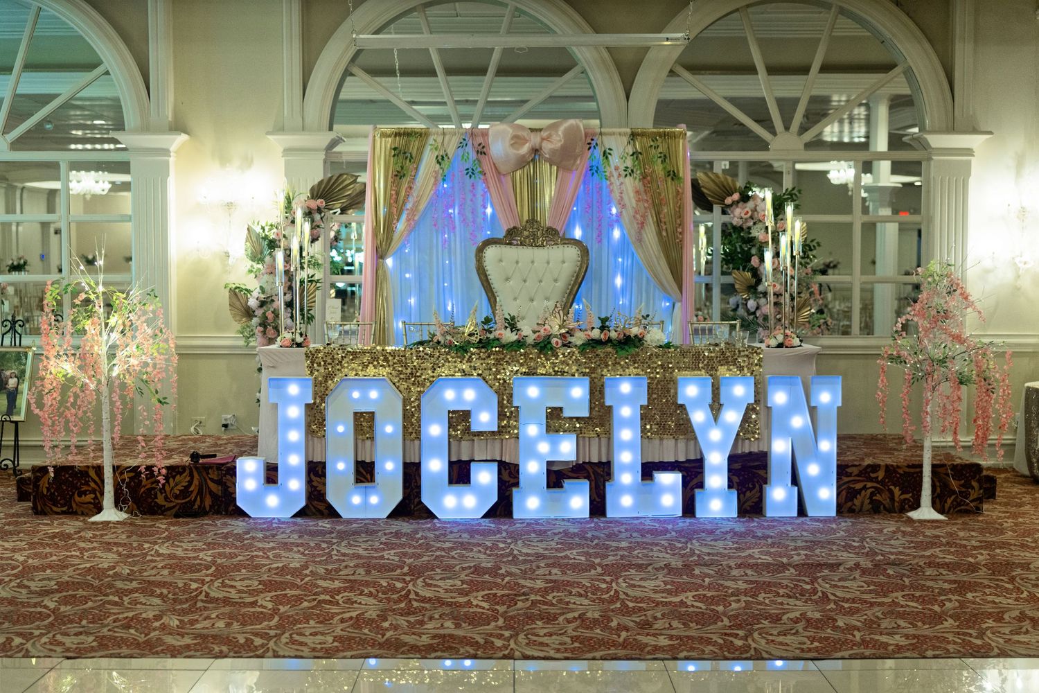 Elegant quinceañera ballroom setup featuring illuminated marquee letters spelling JOCELYN with floral arrangements and decorative backdrop.