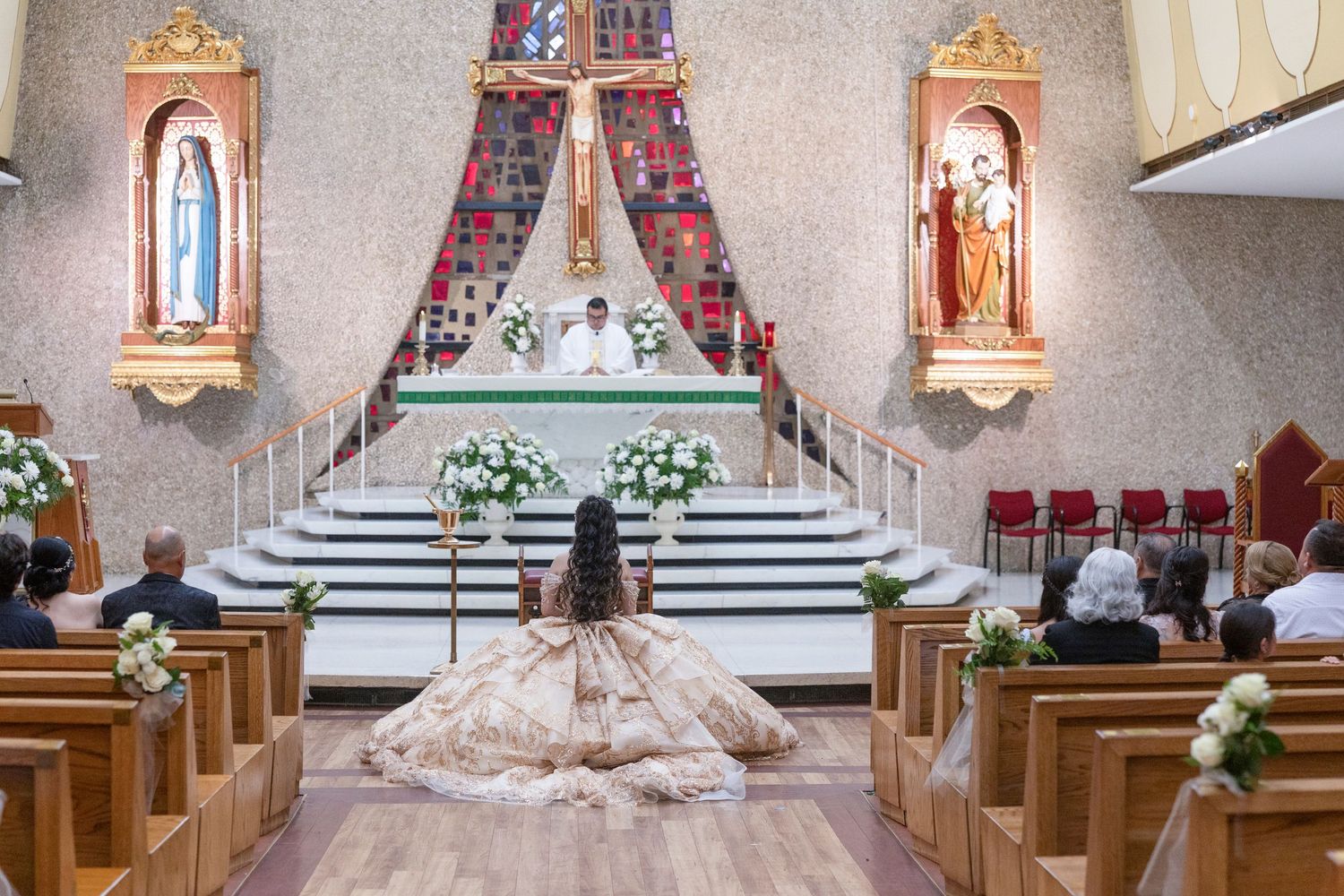 Young woman in a ballgown kneels before a church altar during a quinceañera ceremony with guests seated in pews.