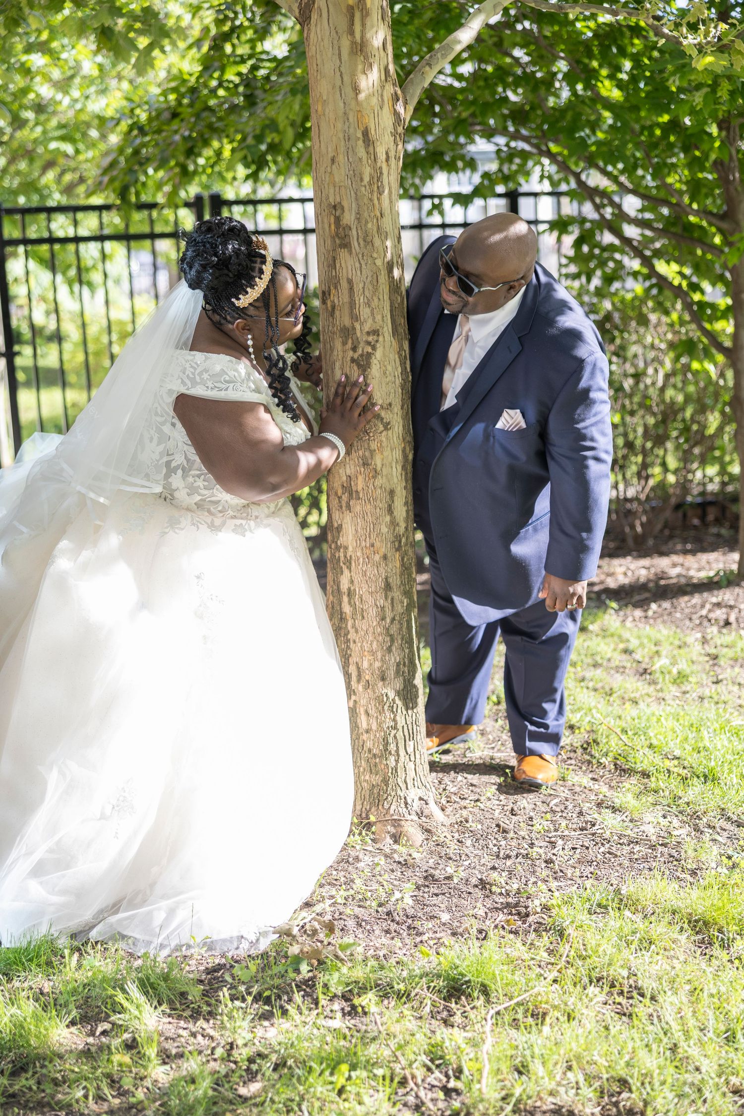 Bride in white ball gown and groom in navy suit share a romantic moment by a tree outdoors.