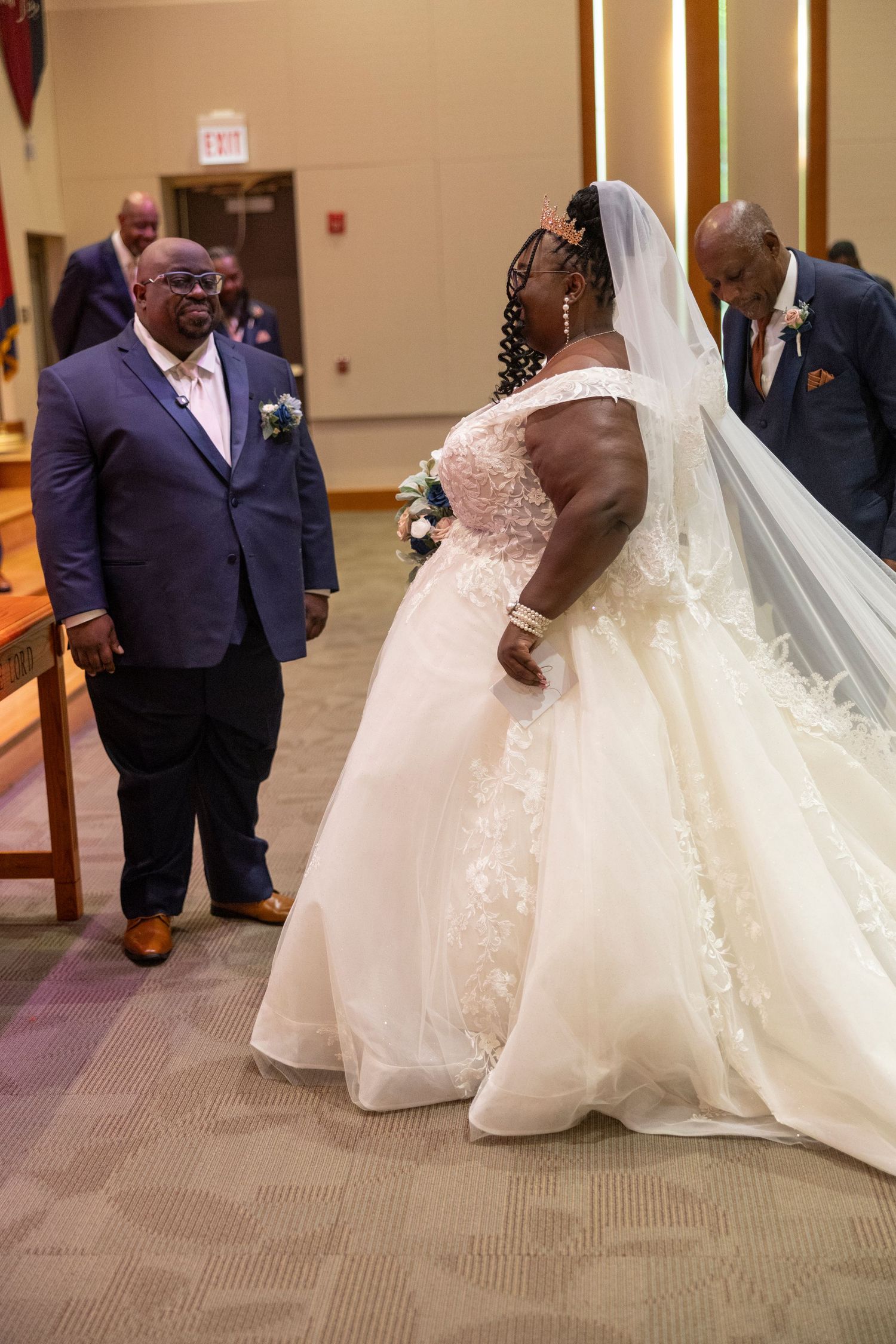 Bride in white lace gown and veil walks down church aisle with groom and groomsmen in blue suits.