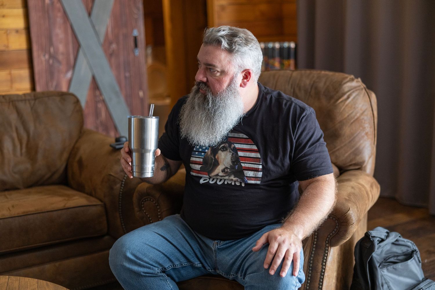 Bearded man with gray beard sits in leather chair holding stainless steel tumbler, wearing American flag t-shirt indoors.
