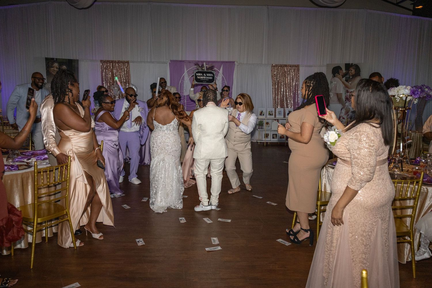 Wedding reception guests dancing in elegant ballroom with gold chairs, purple decor, and bride in white gown.