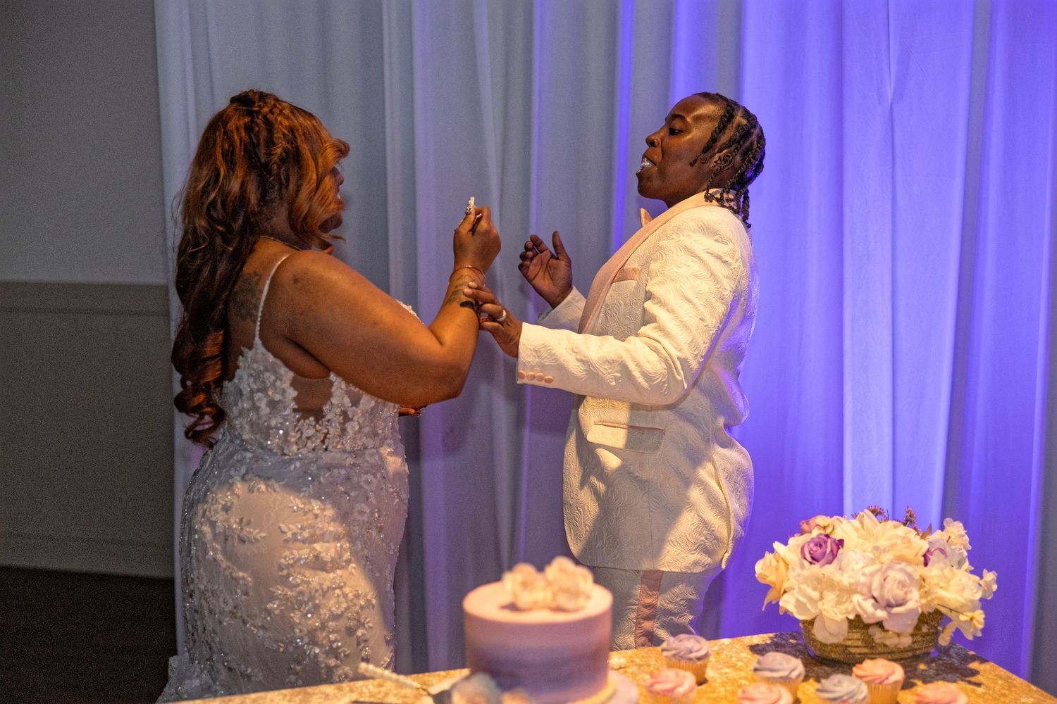 Two brides share a joyful moment at their wedding reception near a decorated cake table with floral arrangements.