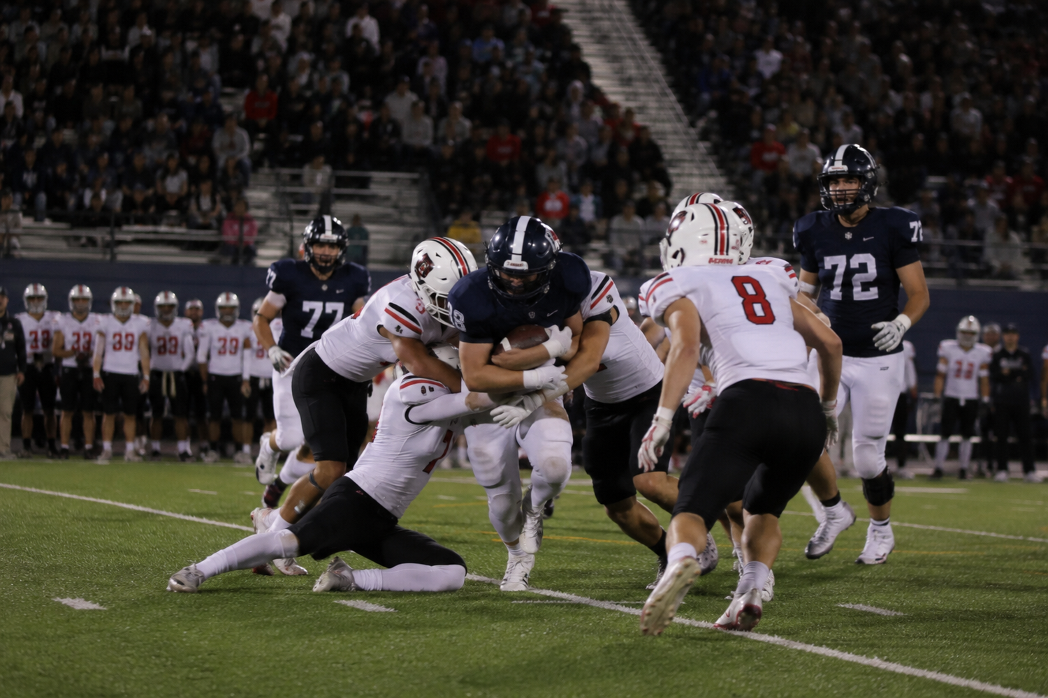 Football players tackle a ball carrier during an intense night game under stadium lights.