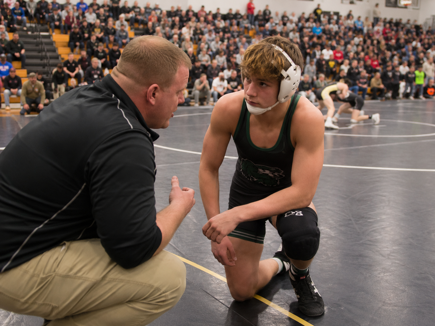 Wrestling coach crouches to give instructions to athlete in singlet during competitive match in crowded gymnasium.