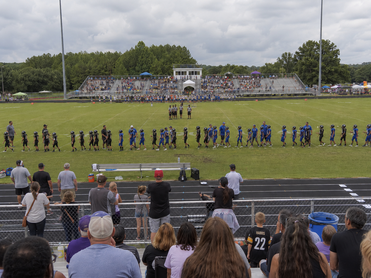Soccer teams line up on a green field before a match as fans watch from bleachers outdoors.