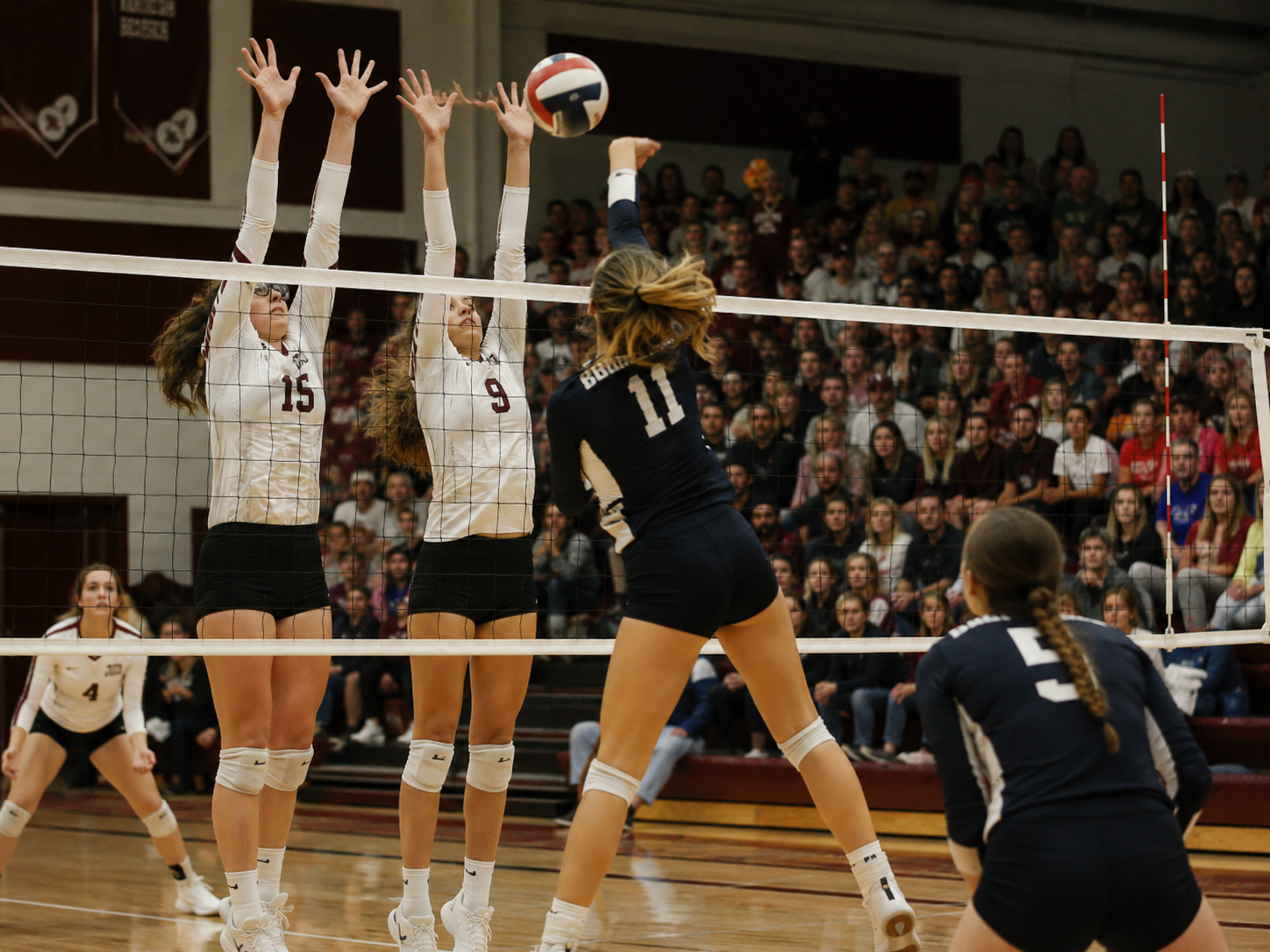 Female volleyball player spikes ball over net as two opponents attempt a block in packed gymnasium.