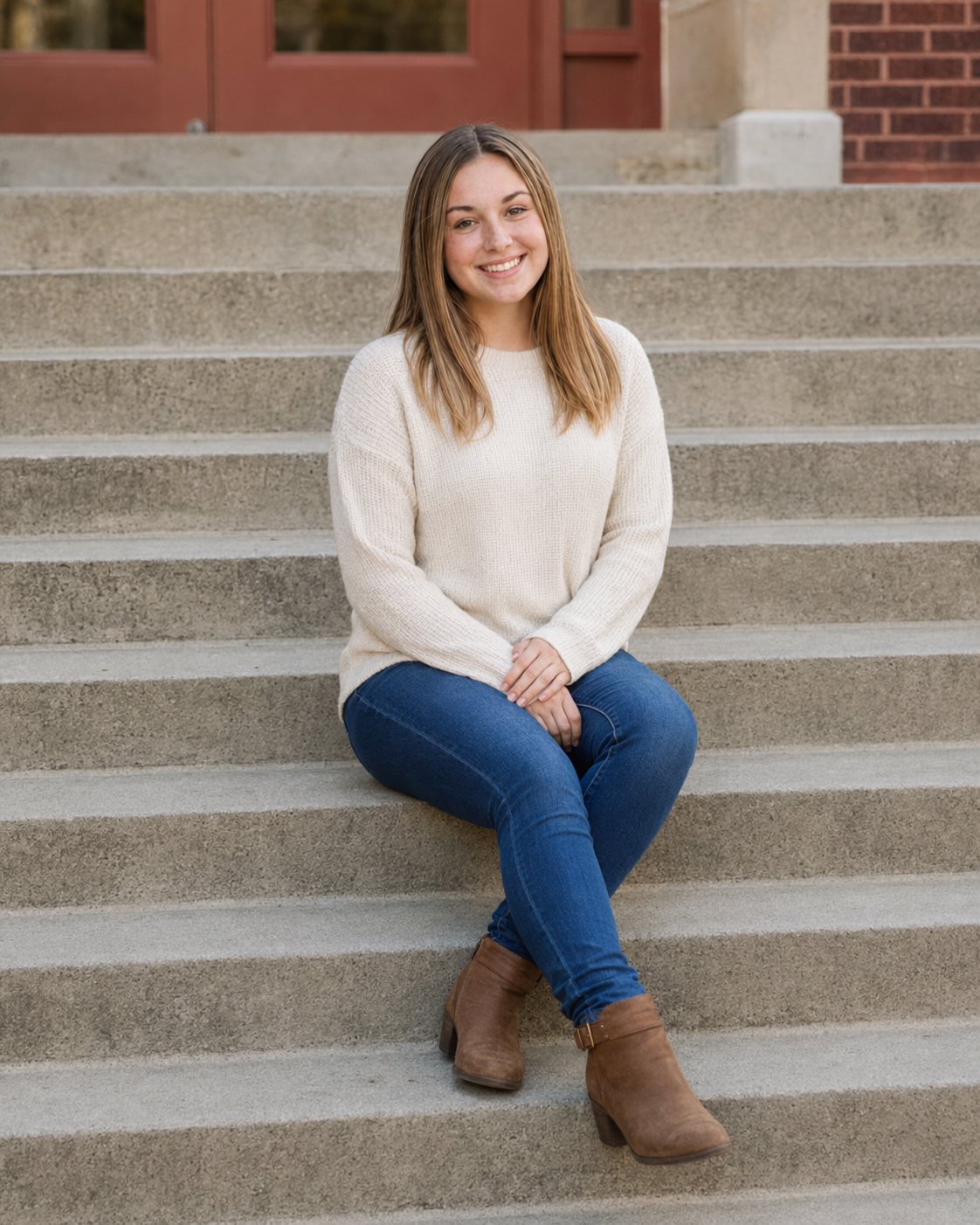 Young woman smiling on concrete steps wearing cream sweater, blue jeans, and brown ankle boots.
