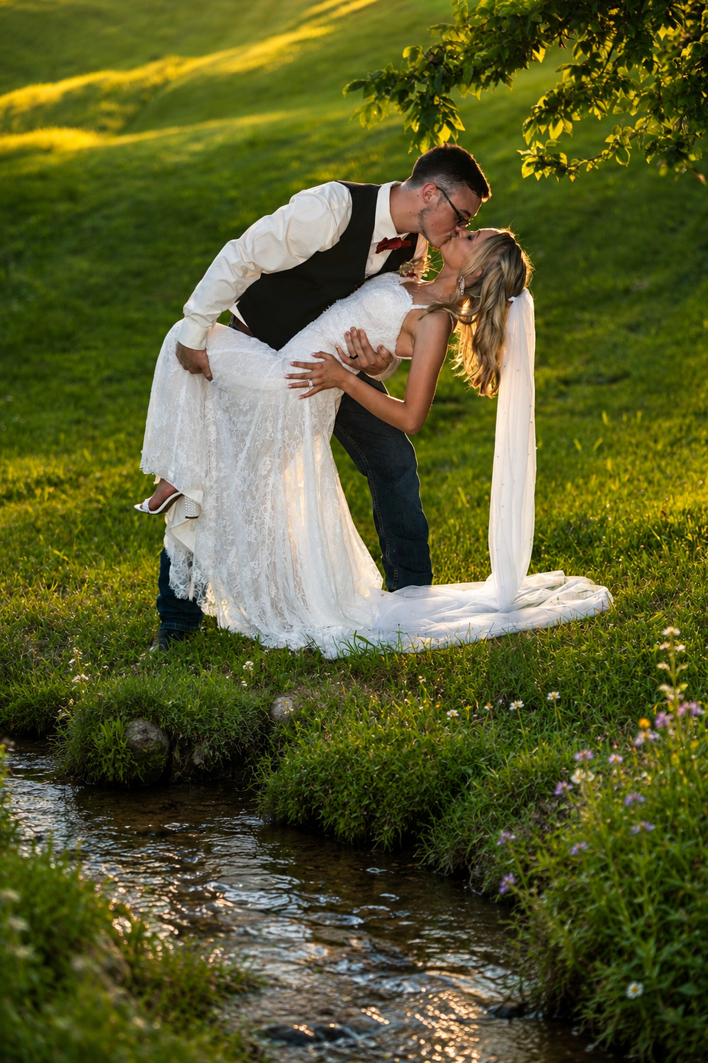 Groom dips bride for a romantic kiss beside a babbling brook in a lush green meadow during golden hour.