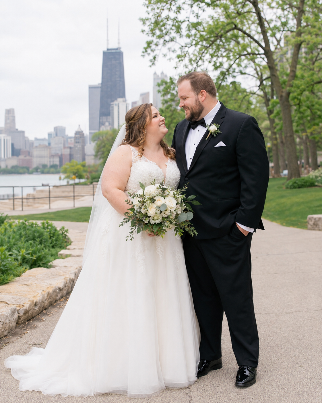 Bride and groom smiling at each other in Chicago with John Hancock tower skyline, bride holding white floral bouquet.