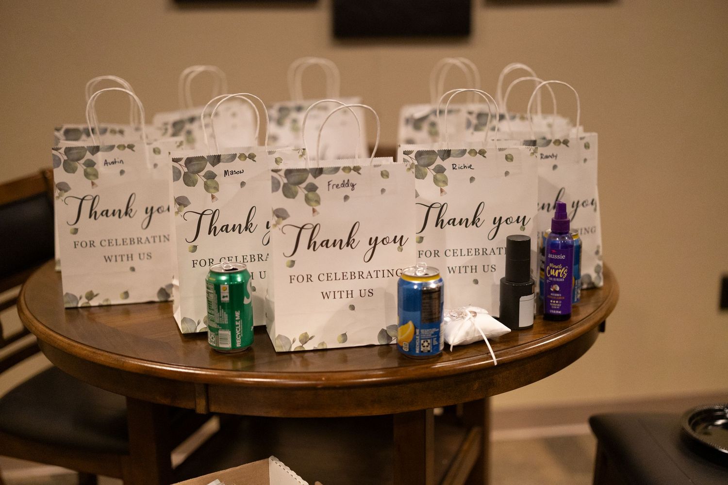 Small white thank you gift bags with cards and colorful mini bottles arranged on a round wooden table.