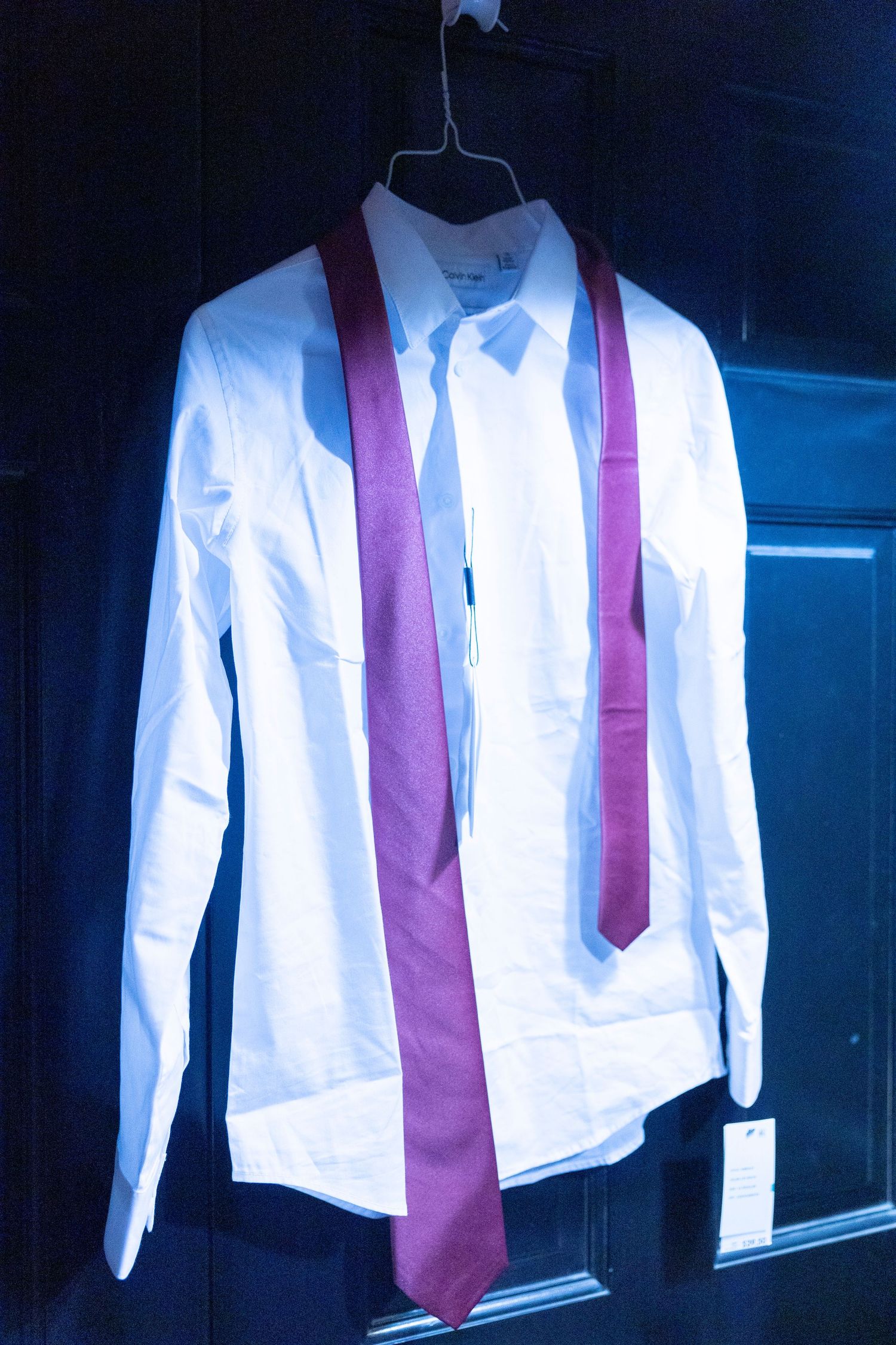 White dress shirt with burgundy necktie hanging on black background in formal attire display.