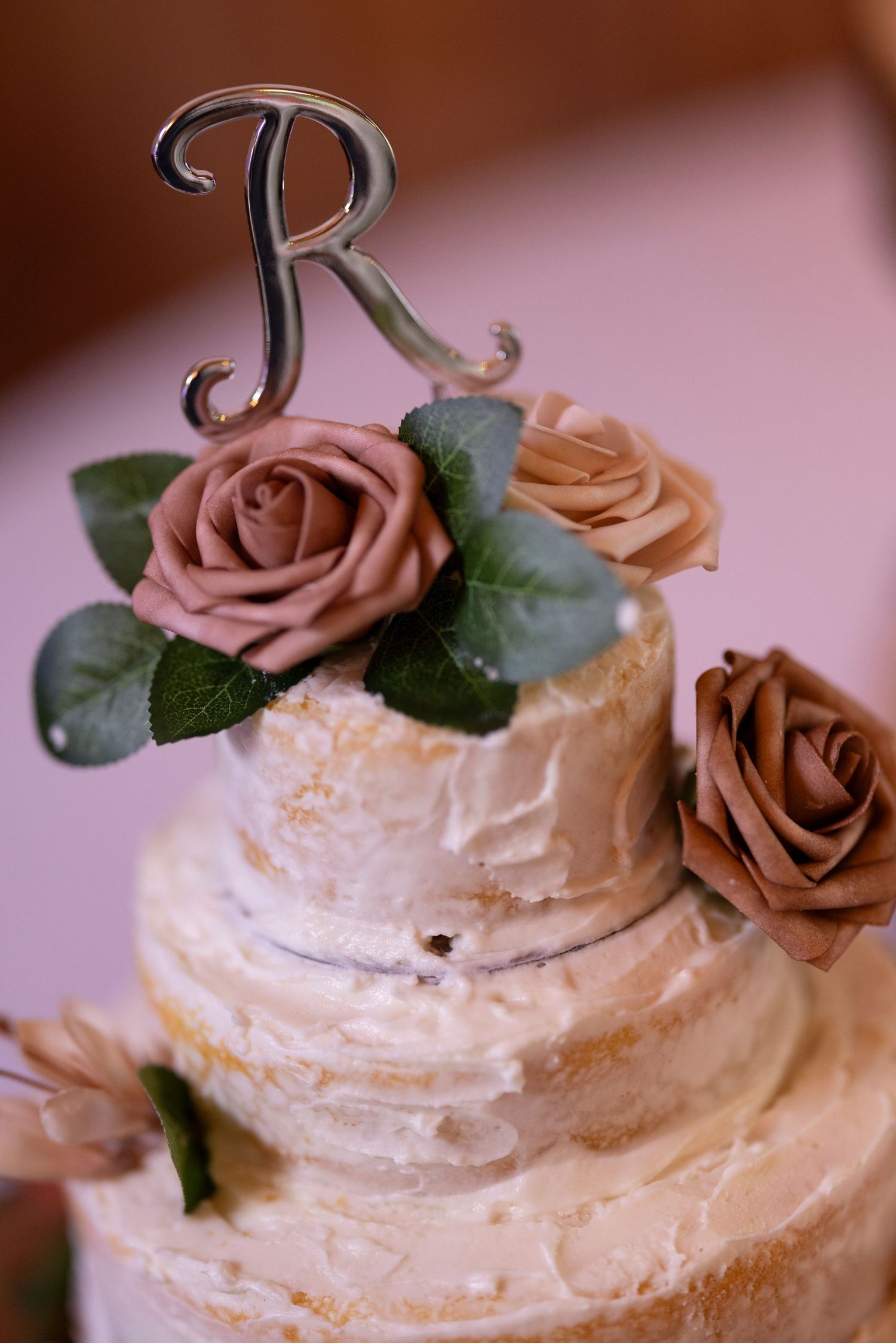 White tiered wedding cake decorated with pink and peach roses and topped with a decorative letter 'R' cake topper.