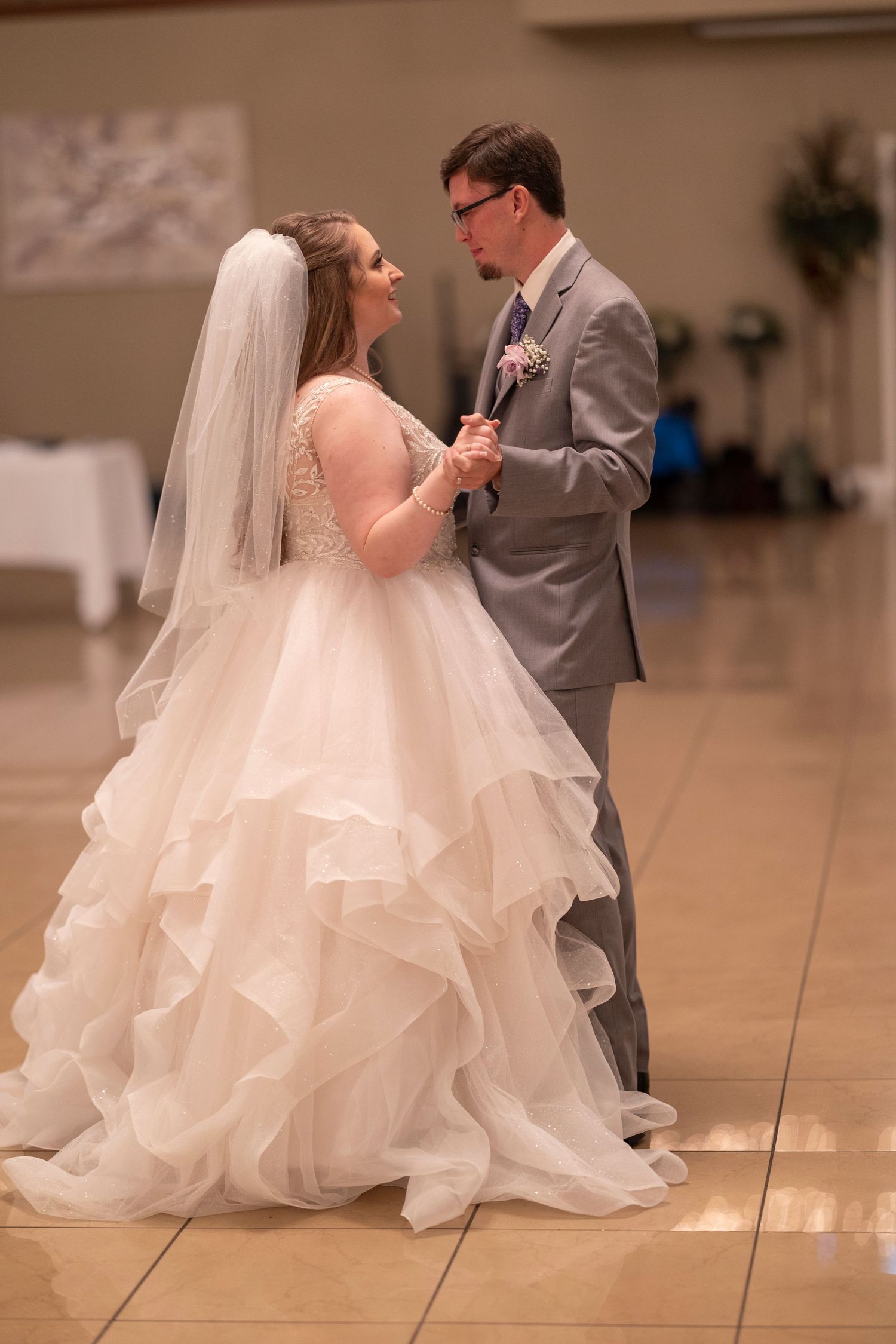 Bride in layered ballgown and groom in gray suit share intimate moment during first dance.