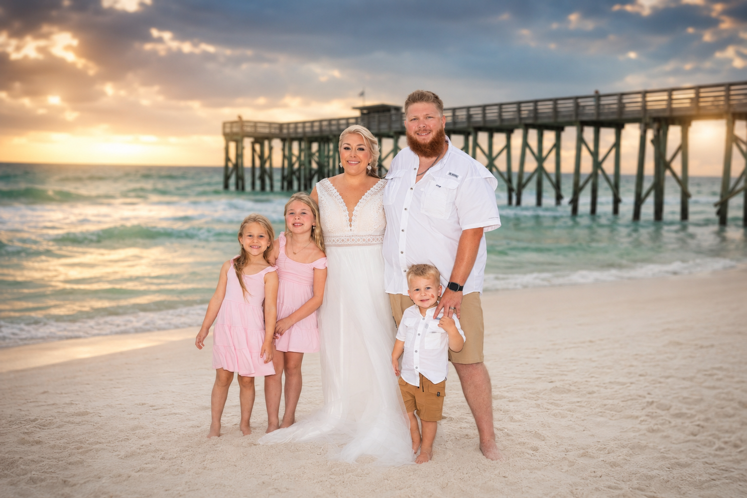 Family of five poses on a sandy beach at sunset near a pier, with parents in wedding attire and children in pink.