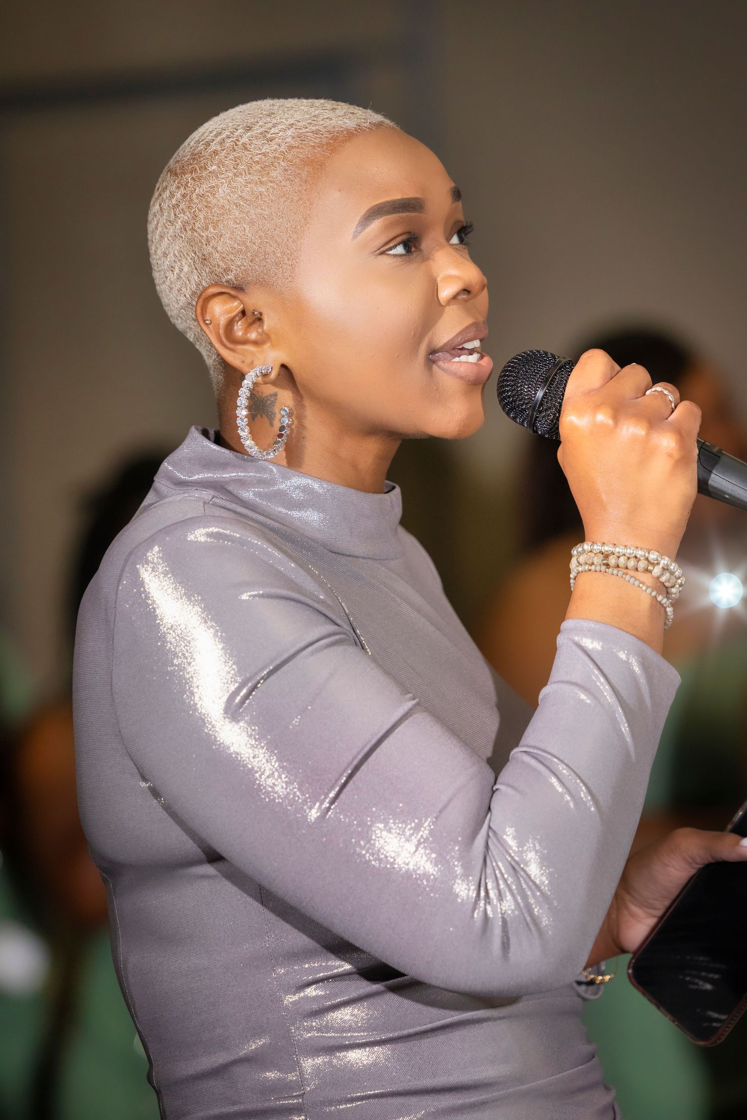 A woman with short blonde hair sings into a microphone, wearing a silver metallic dress and hoop earrings.