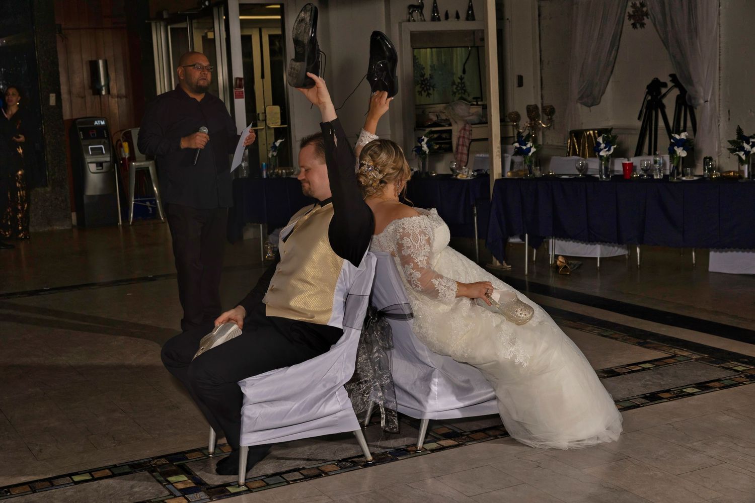 Bride and groom playing wedding shoe game, holding shoes up while seated back-to-back on dance floor at reception.