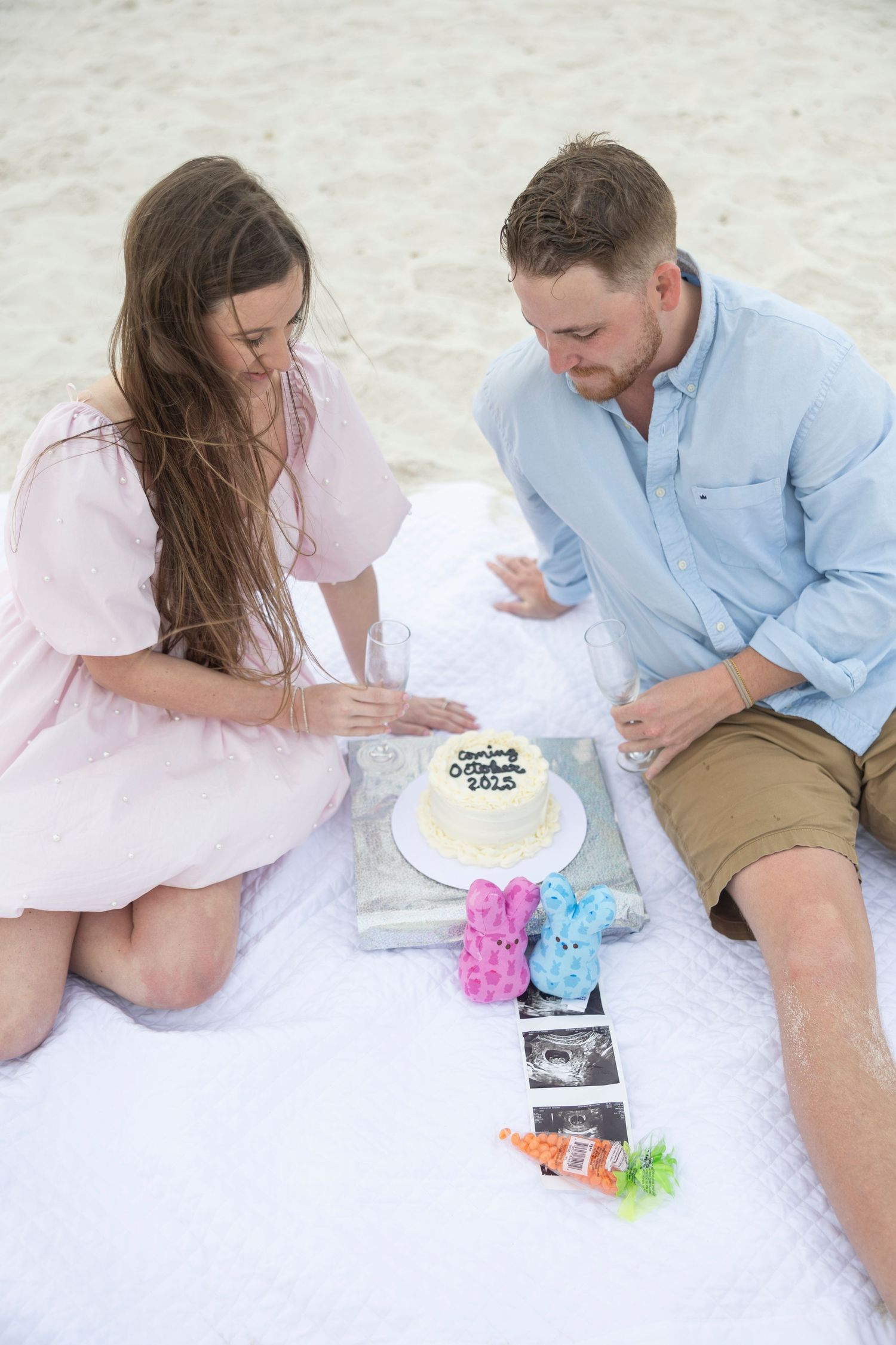 Couple sitting on beach blanket with pregnancy announcement cake, ultrasound photos, and baby booties on white sand.