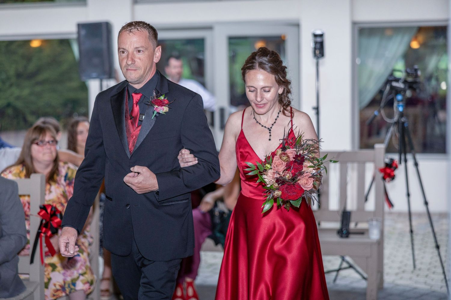 A couple in formal red and black attire walk arm in arm during an evening wedding ceremony.