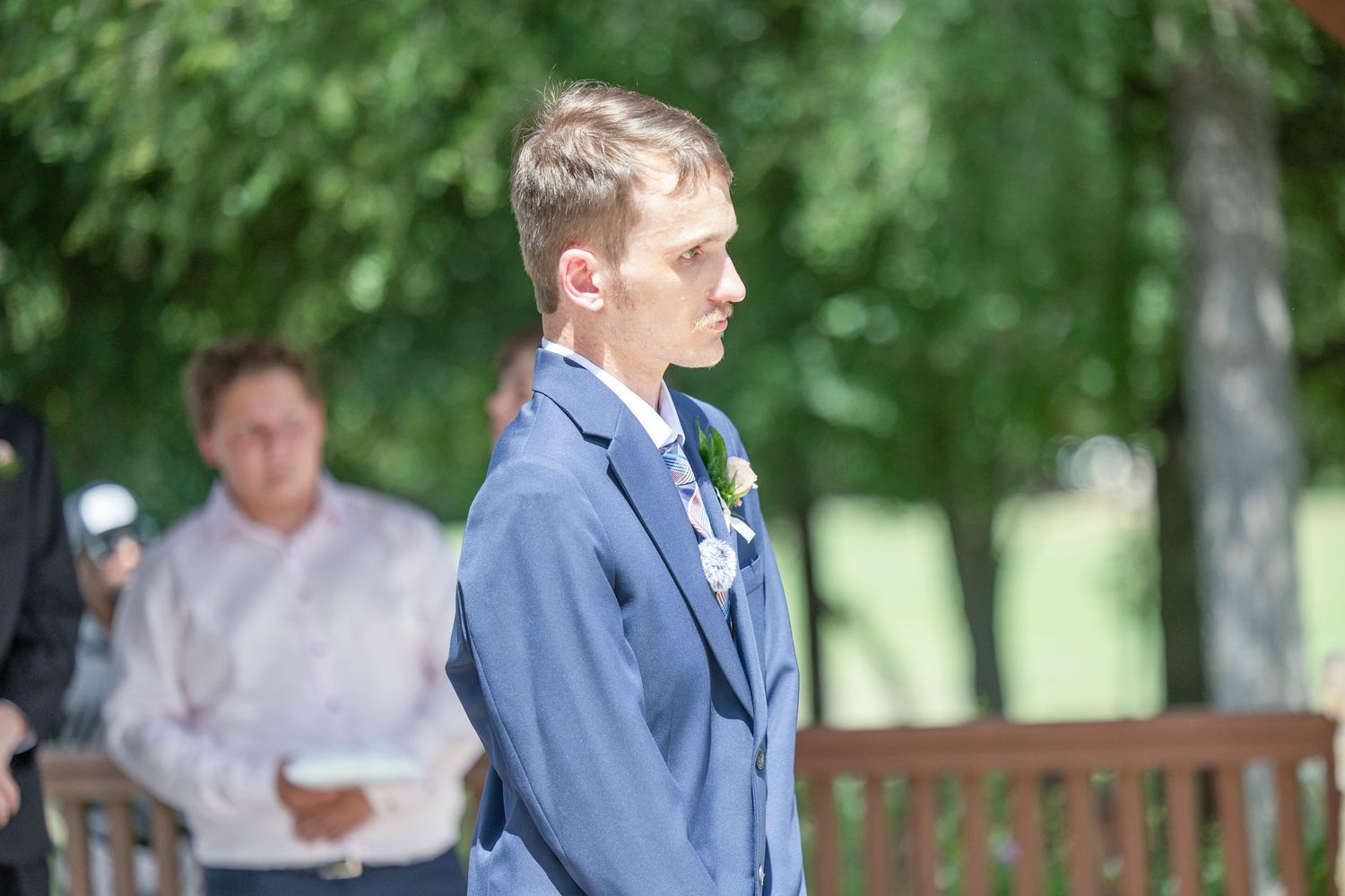 Wedding party gathers outdoors in a garden setting with groomsmen in blue suits and bridesmaids in light dresses.
