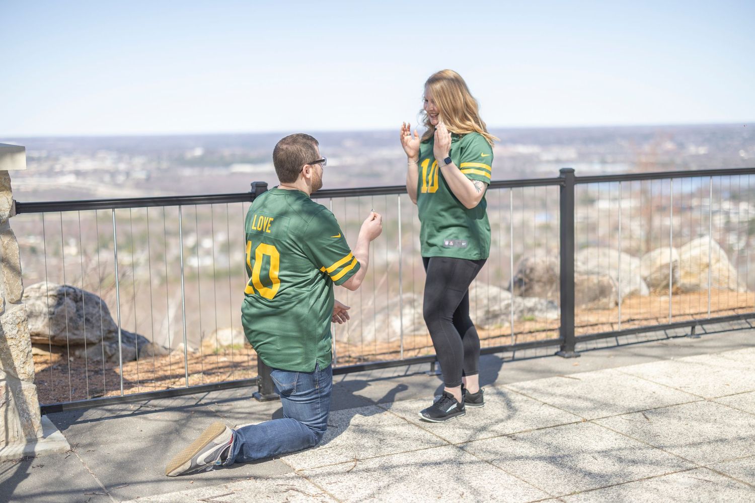 People in green backpacks sit along a stone wall overlooking a scenic mountain valley vista on a sunny day.