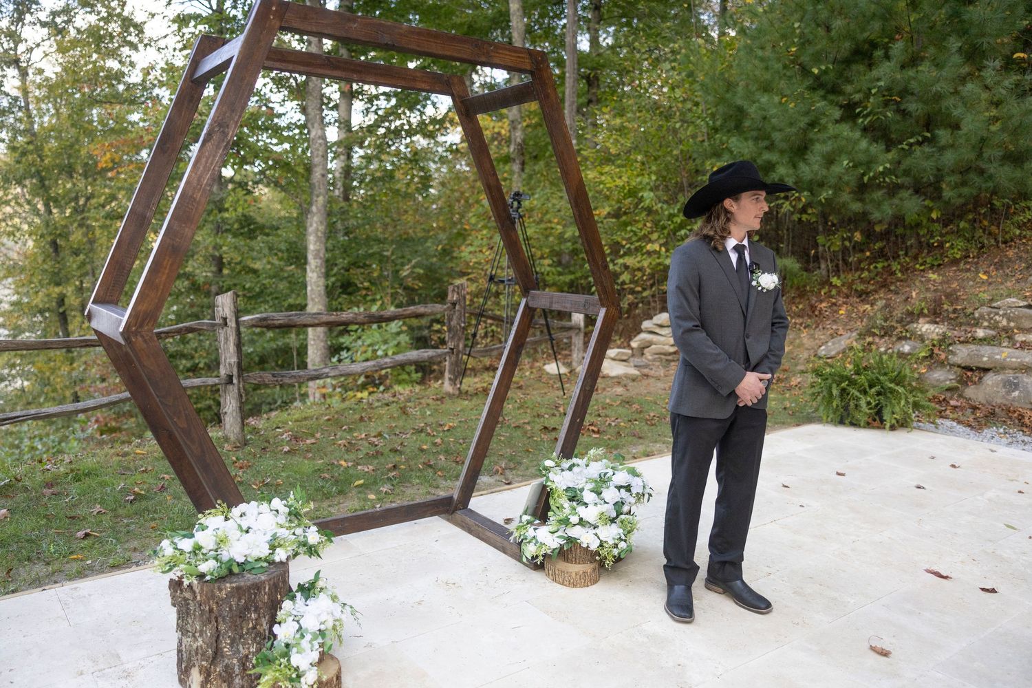A hexagonal wooden arch decorated with white flowers stands on a concrete pad in an outdoor wooded setting.