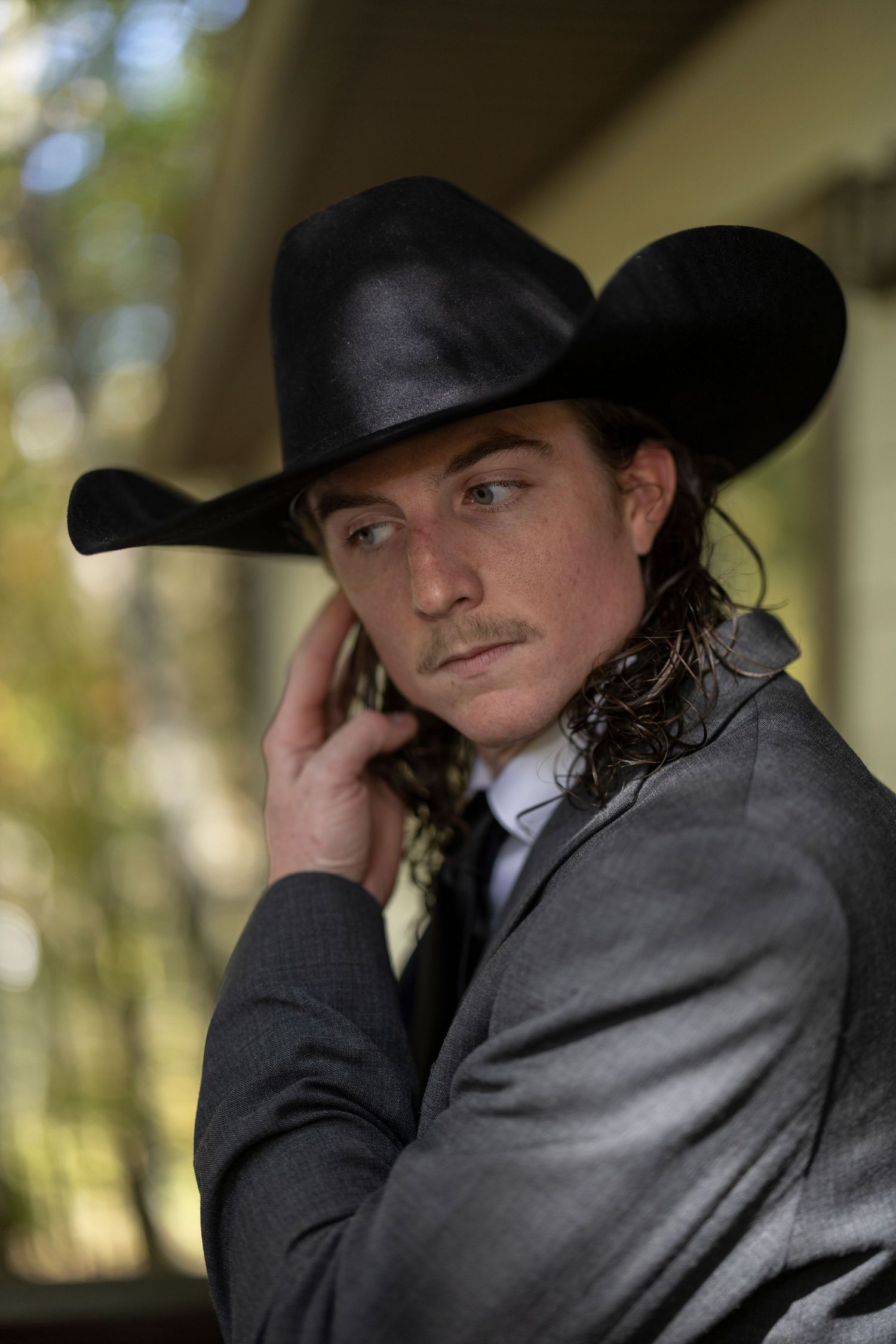 A person in western attire wearing a black cowboy hat and leather jacket poses against a blurred outdoor background.