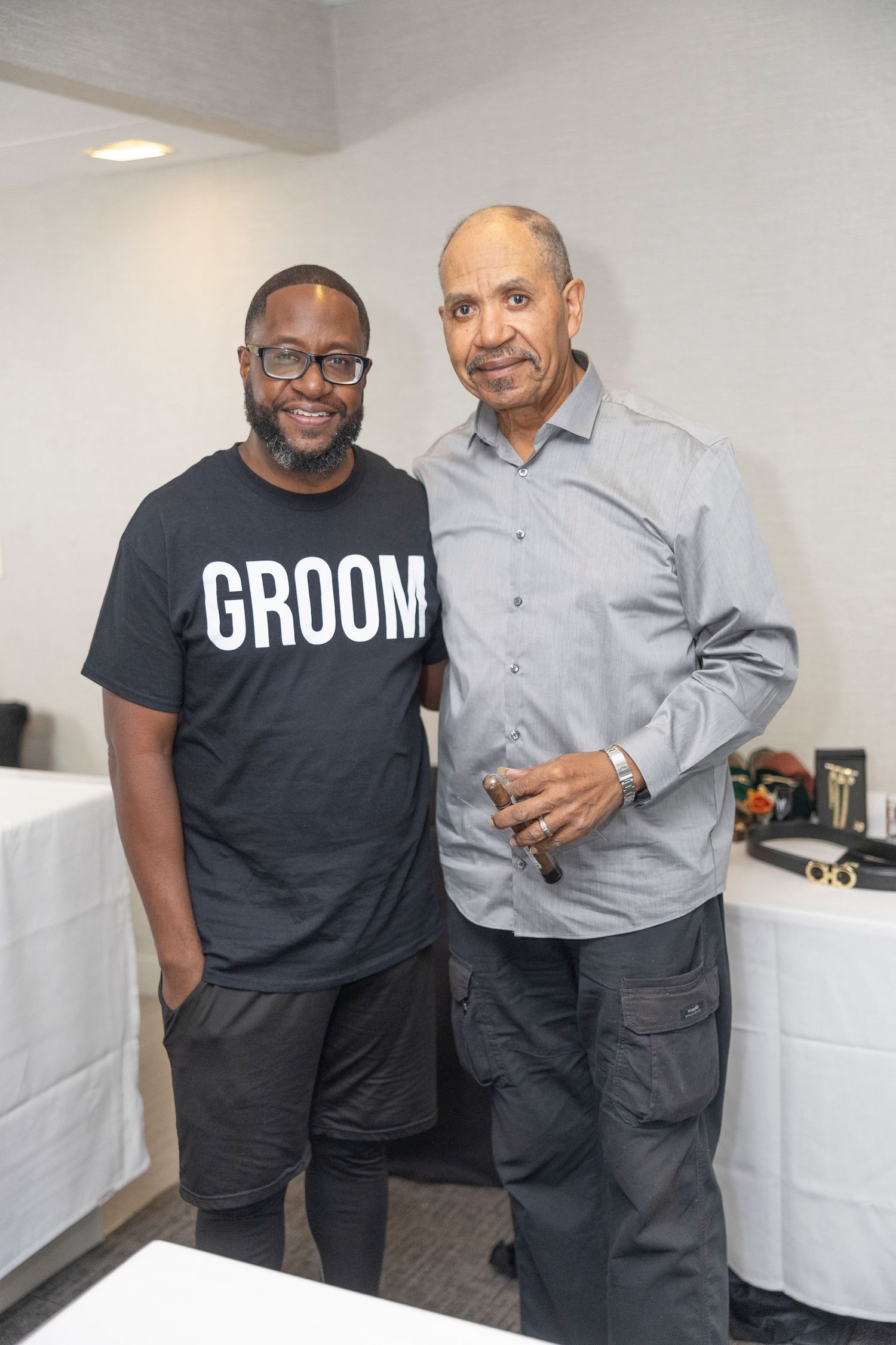 Two men stand together indoors, with one wearing a black 'GROOM' t-shirt and the other in a gray button-down shirt.