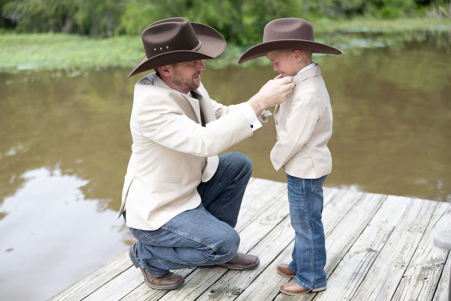 Two people in cowboy hats and jeans sit together on a wooden dock by a peaceful pond.