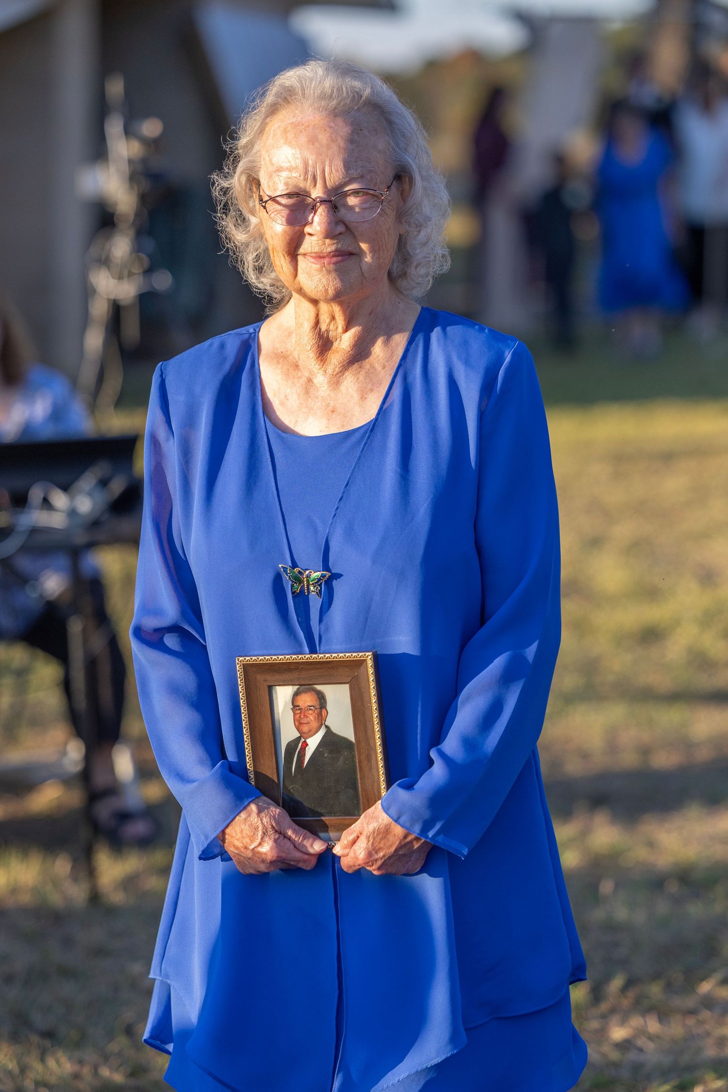 A person in a vibrant royal blue dress holds a framed photograph while standing outdoors in warm sunlight.