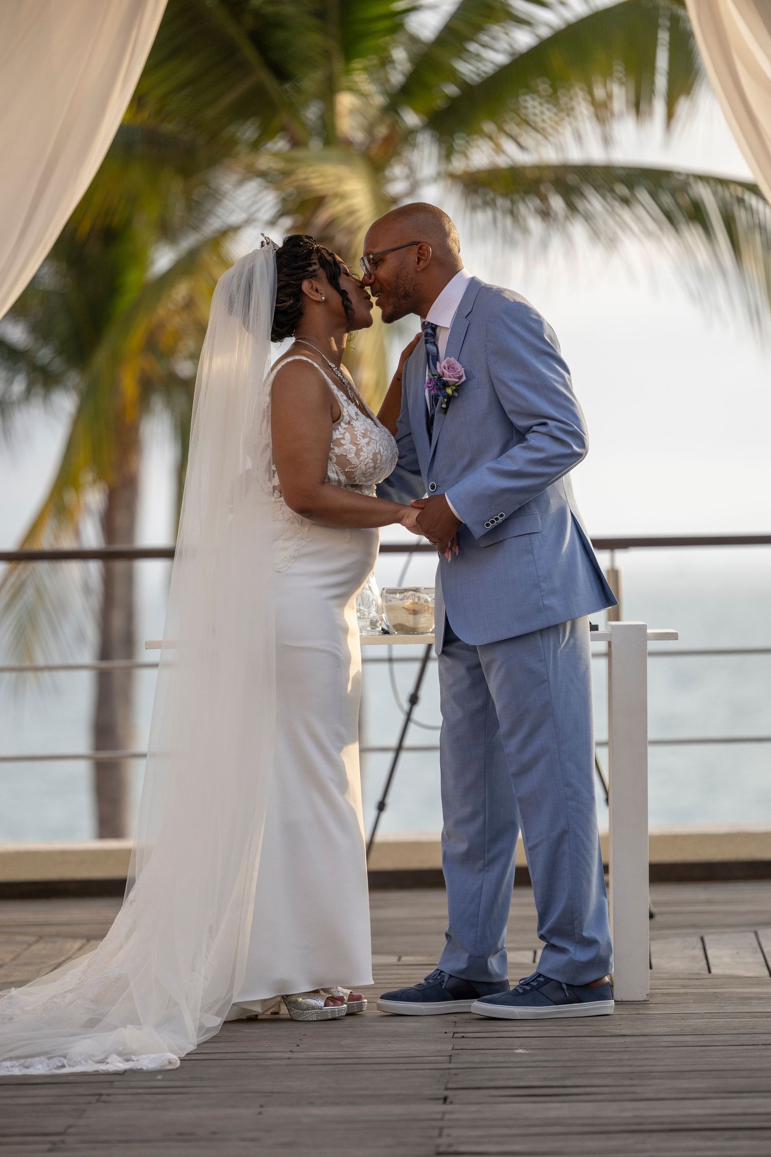 A couple shares a romantic moment on a wooden deck overlooking the ocean with palm trees in the background.