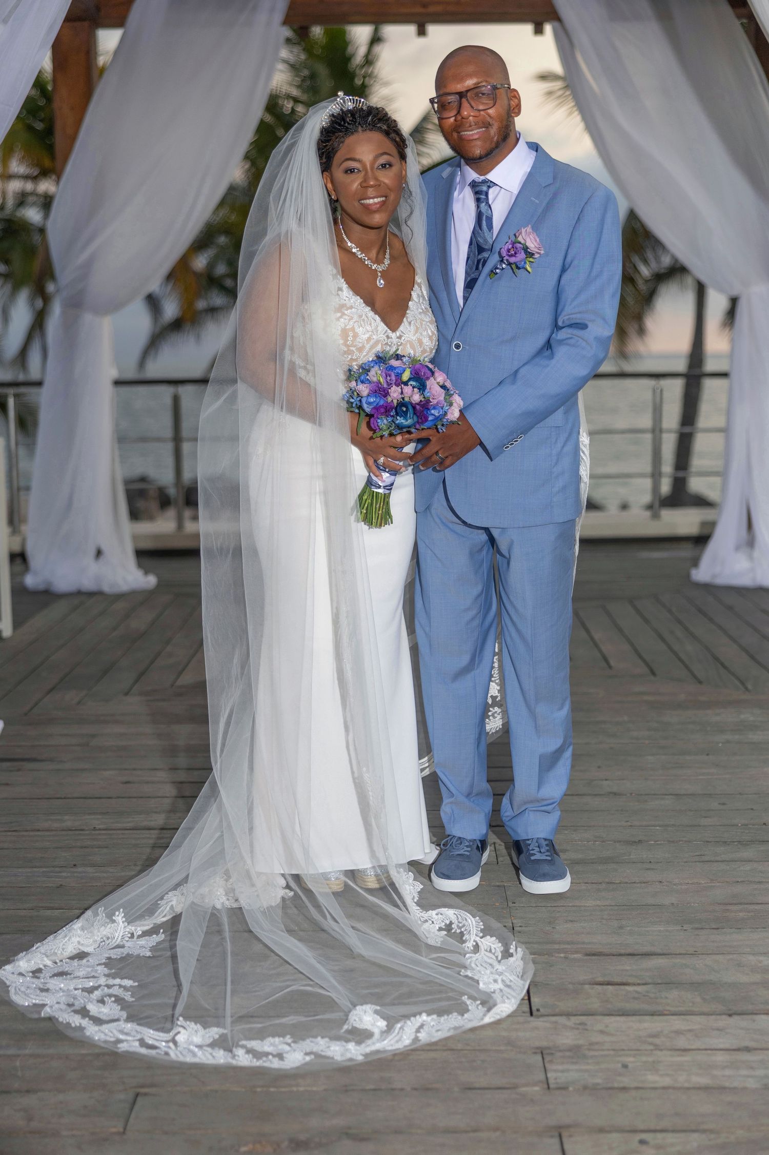 A wedding couple stands together on a wooden deck with white drapery and decorations visible in the background.
