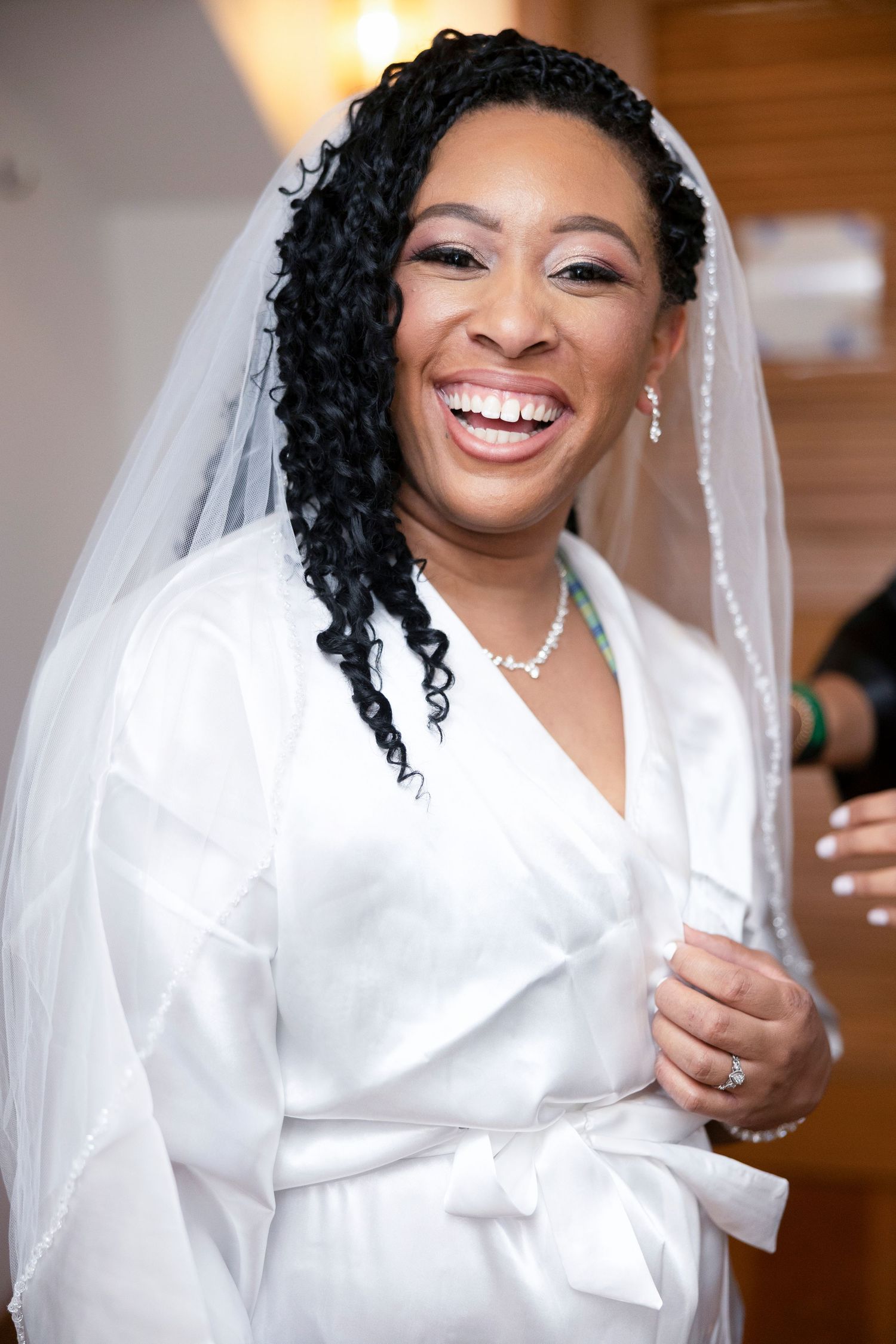 A radiant bride wearing a white satin robe and veil smiles joyfully while getting ready on her wedding day.