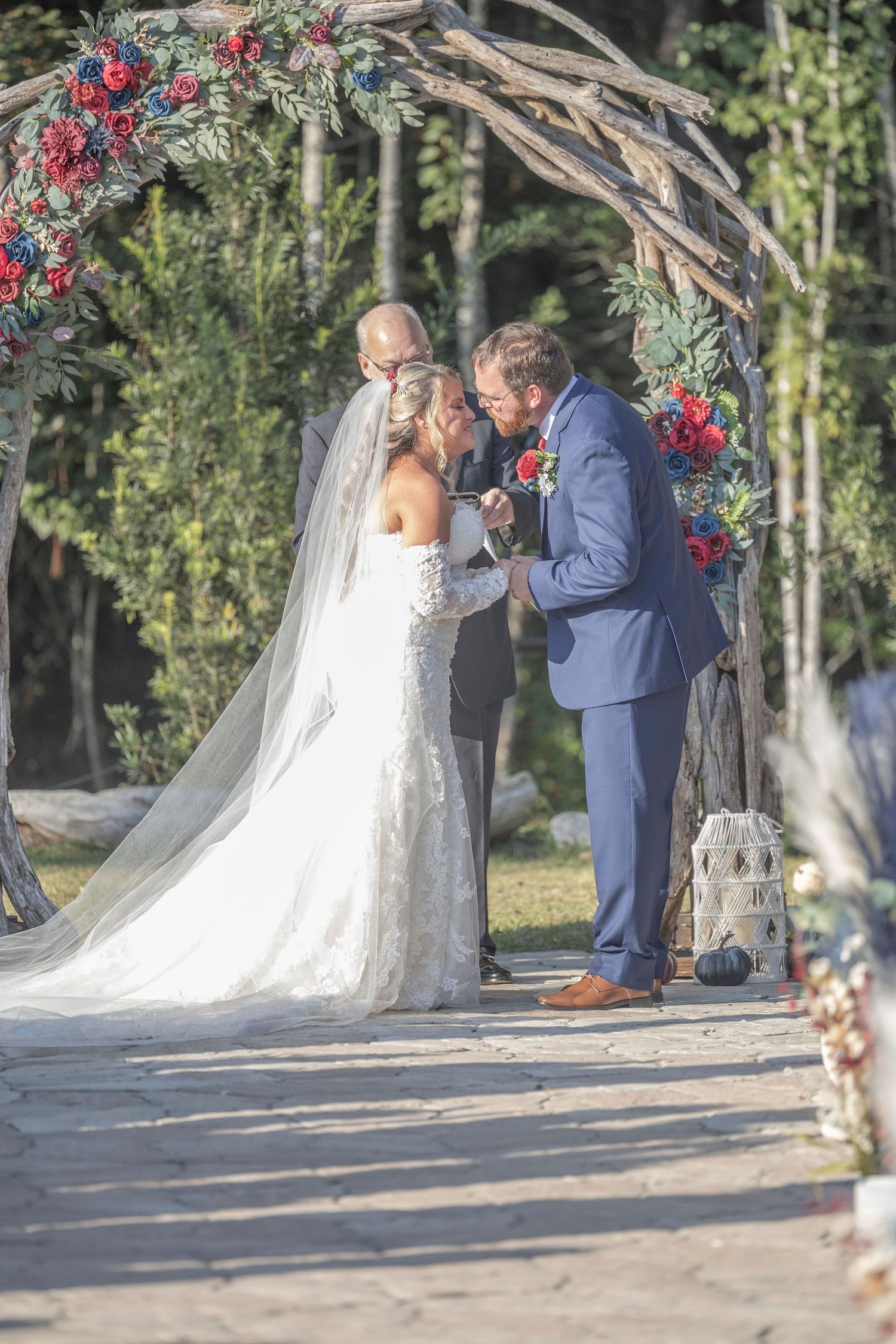 A wedding couple shares an intimate moment under a floral arch decorated with red blooms in an outdoor garden setting.