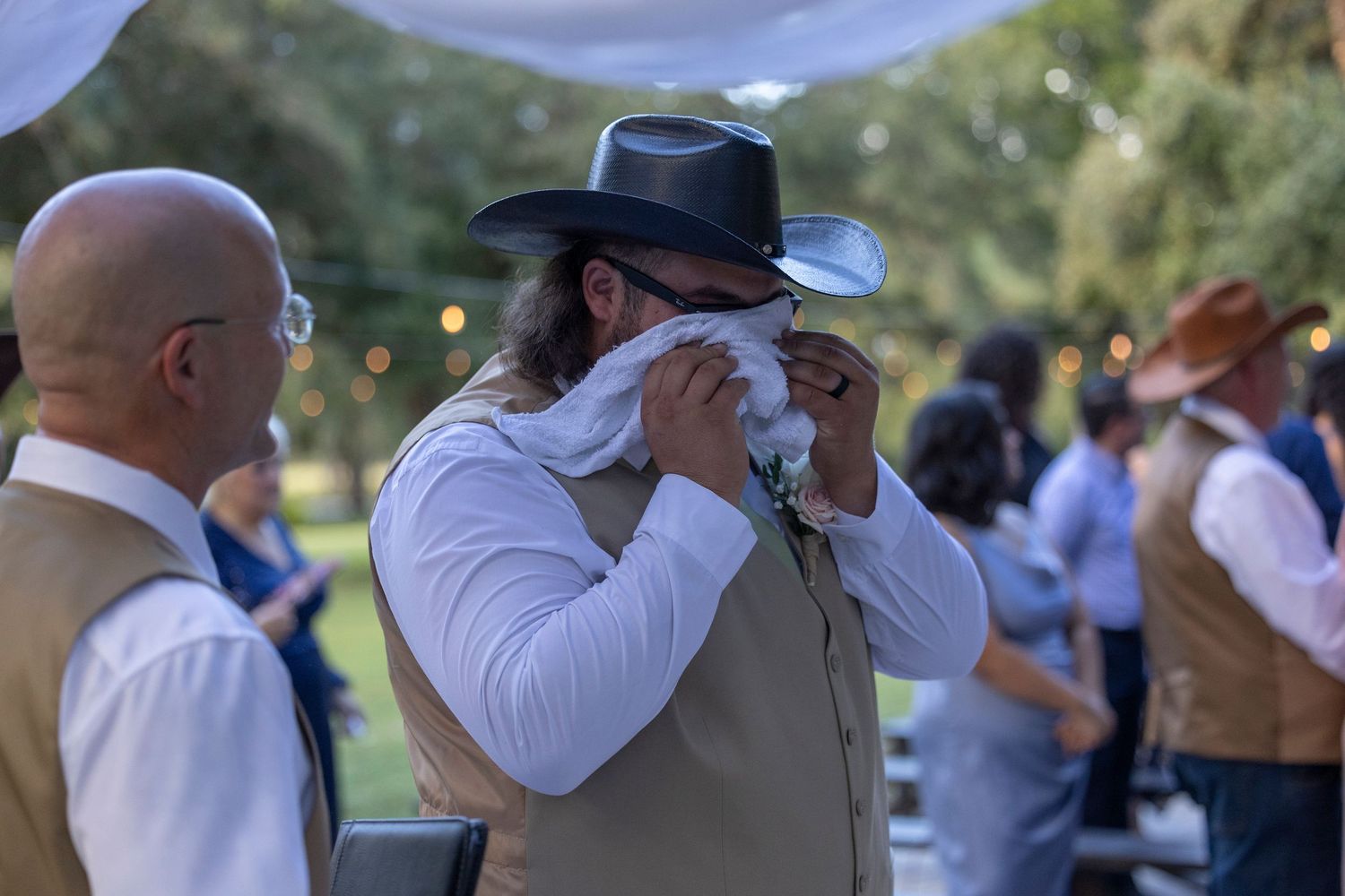 A person in a cowboy hat and tan vest wipes their eyes during an emotional outdoor gathering.