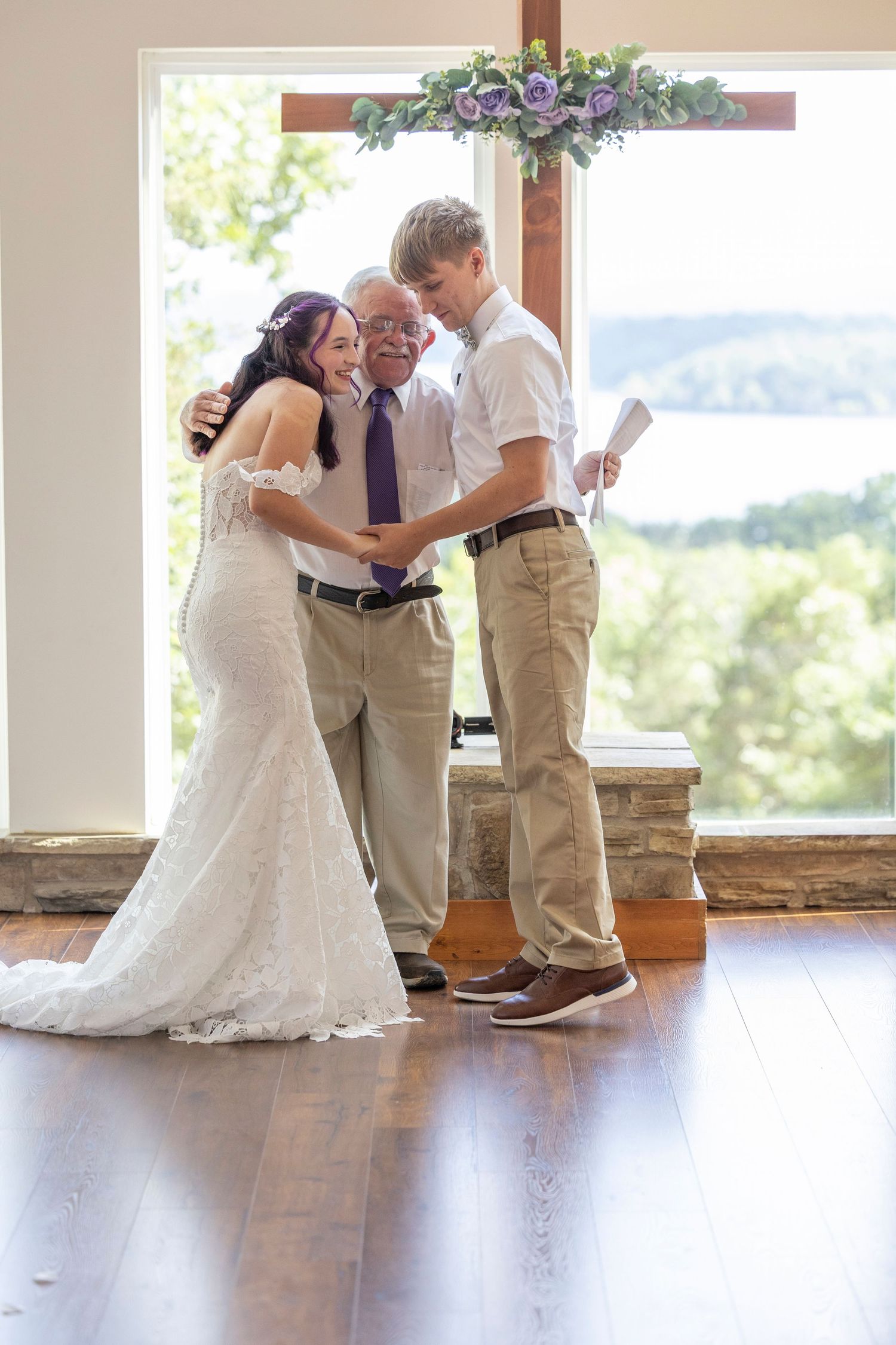 Bride and groom exchange vows with officiant before a wooden cross at an elegant indoor wedding ceremony.
